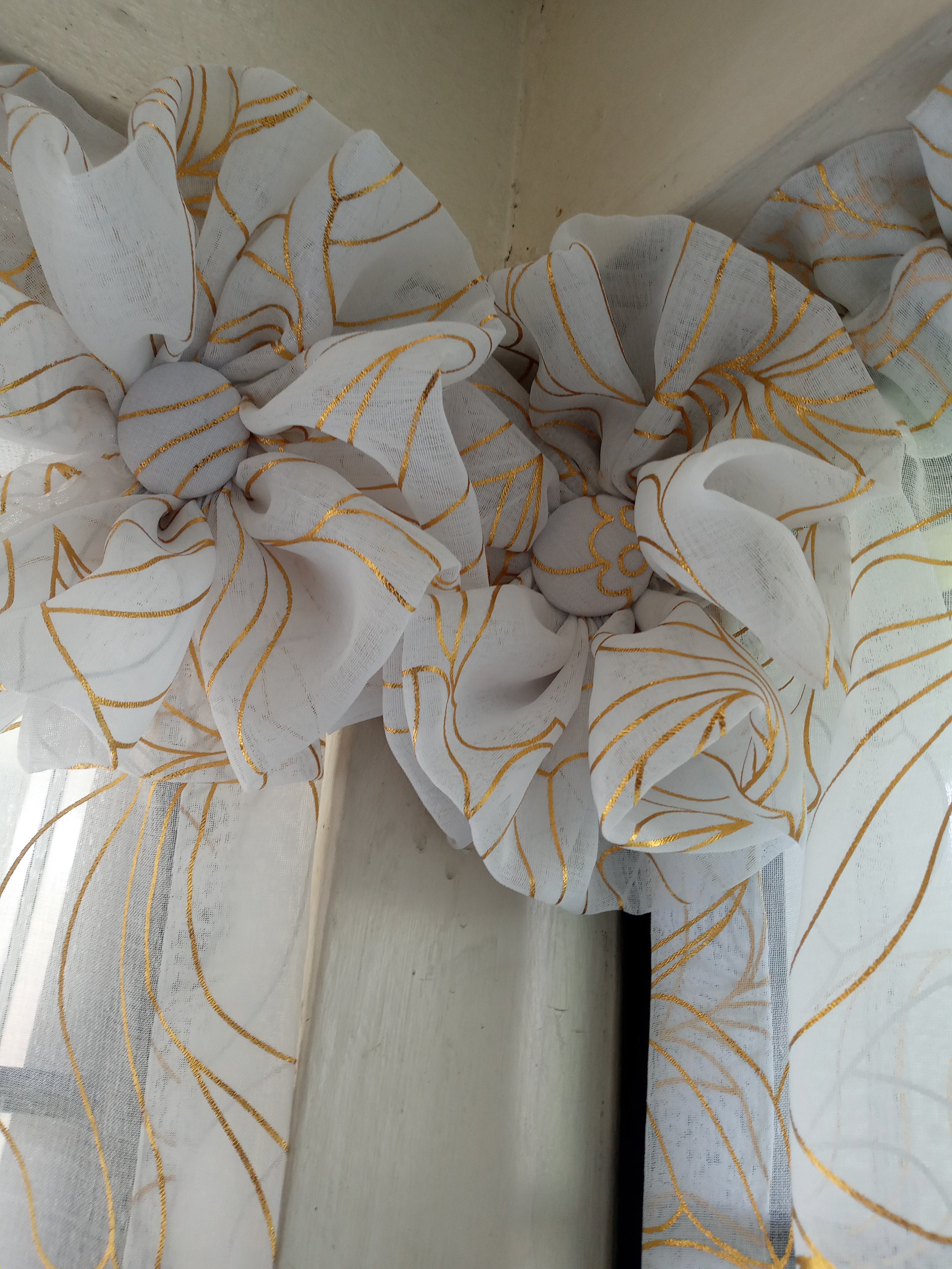 Close-up of a white sheer curtain with gold leaf pattern, tied back with a fabric tie, hanging by a window.