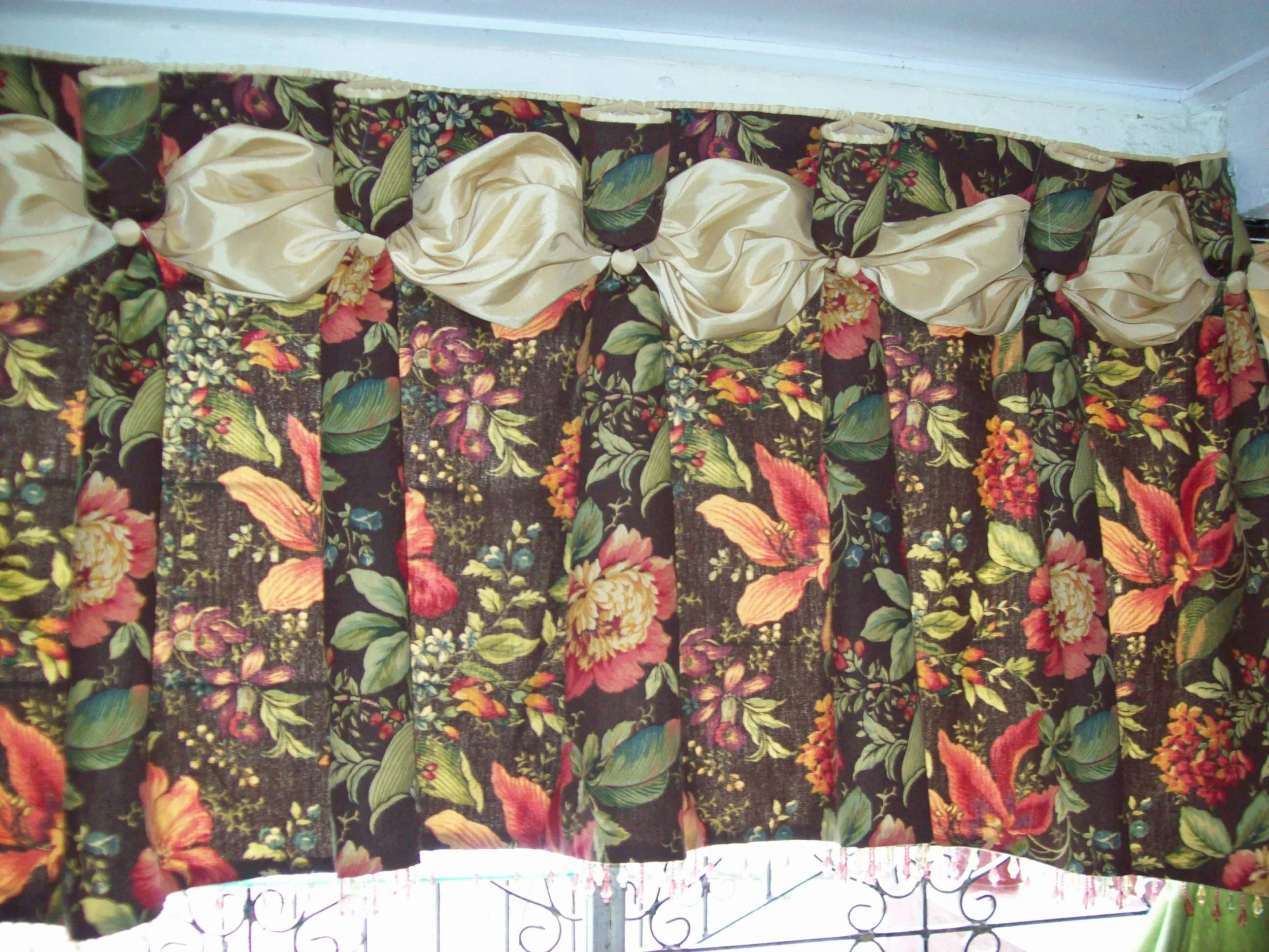 A floral-patterned curtain with large flowers and leaves in pink, yellow, green, and blue on a dark background, topped with cream-colored satin bows with white pom-pom accents.