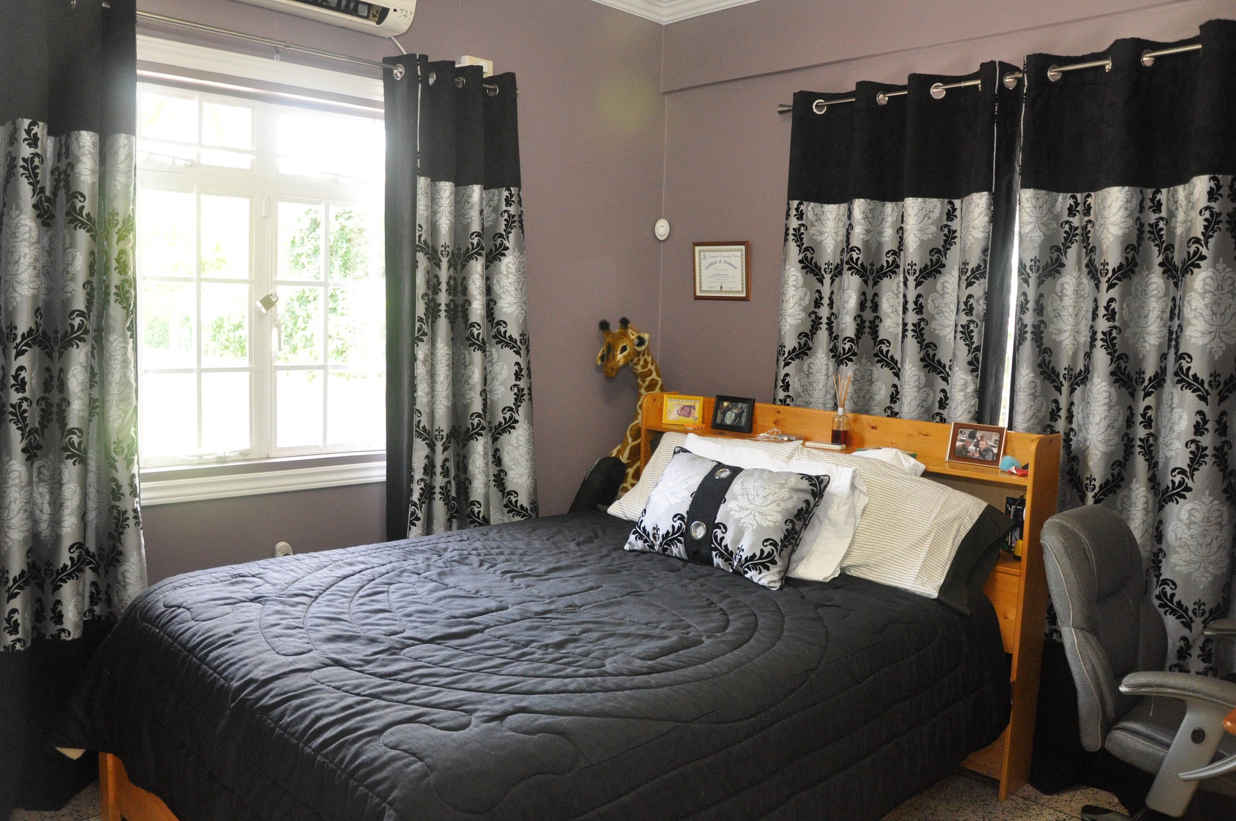 Bedroom with a bed covered in a black quilt, black and white decor, a window with black and white curtains, a plush giraffe toy, framed photos, a reading chair, and a purple wall.