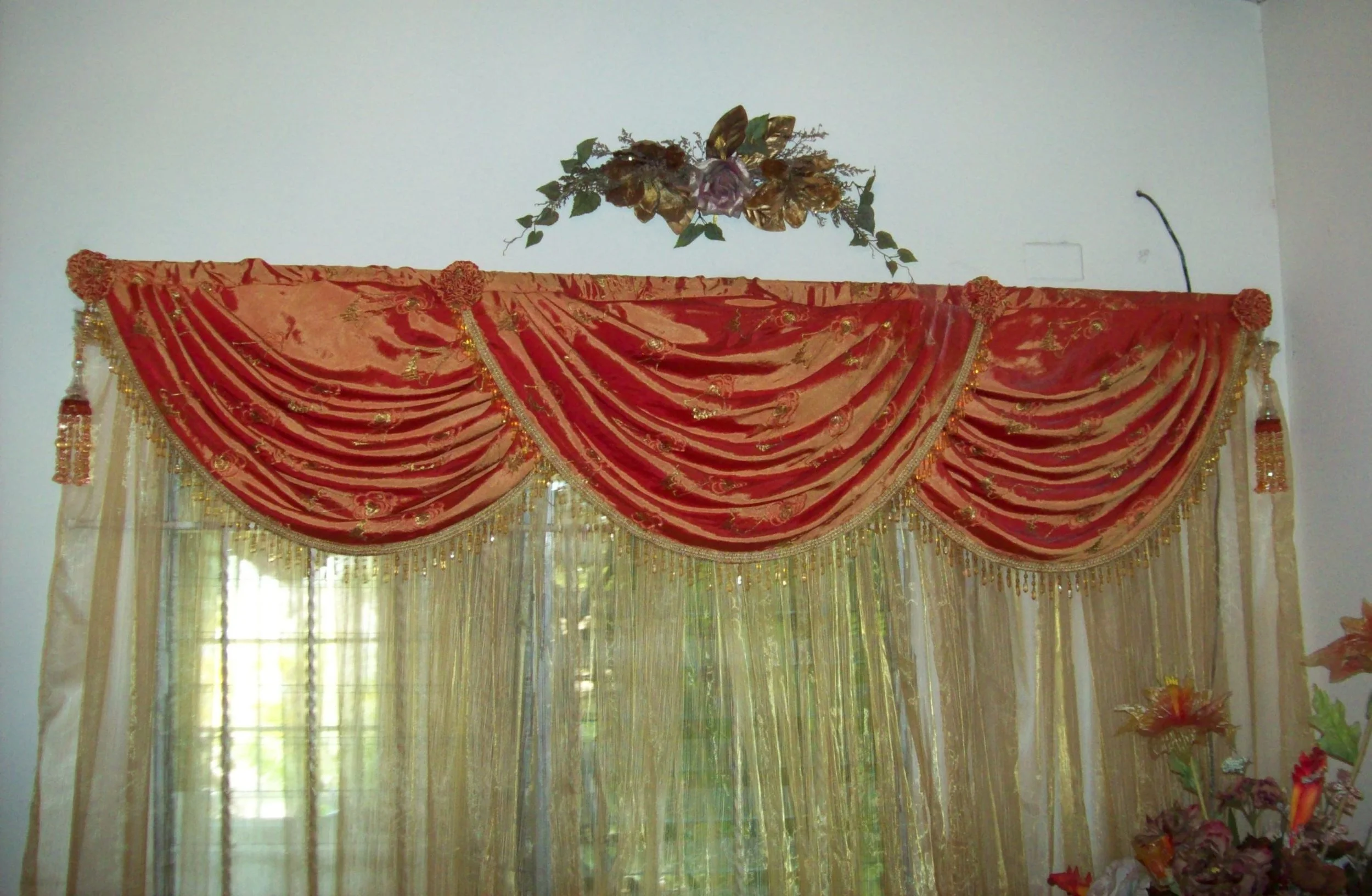 Decorative red and gold curtain with embroidered patterns and tassels hanging on a window, with a floral arrangement above it on the wall.