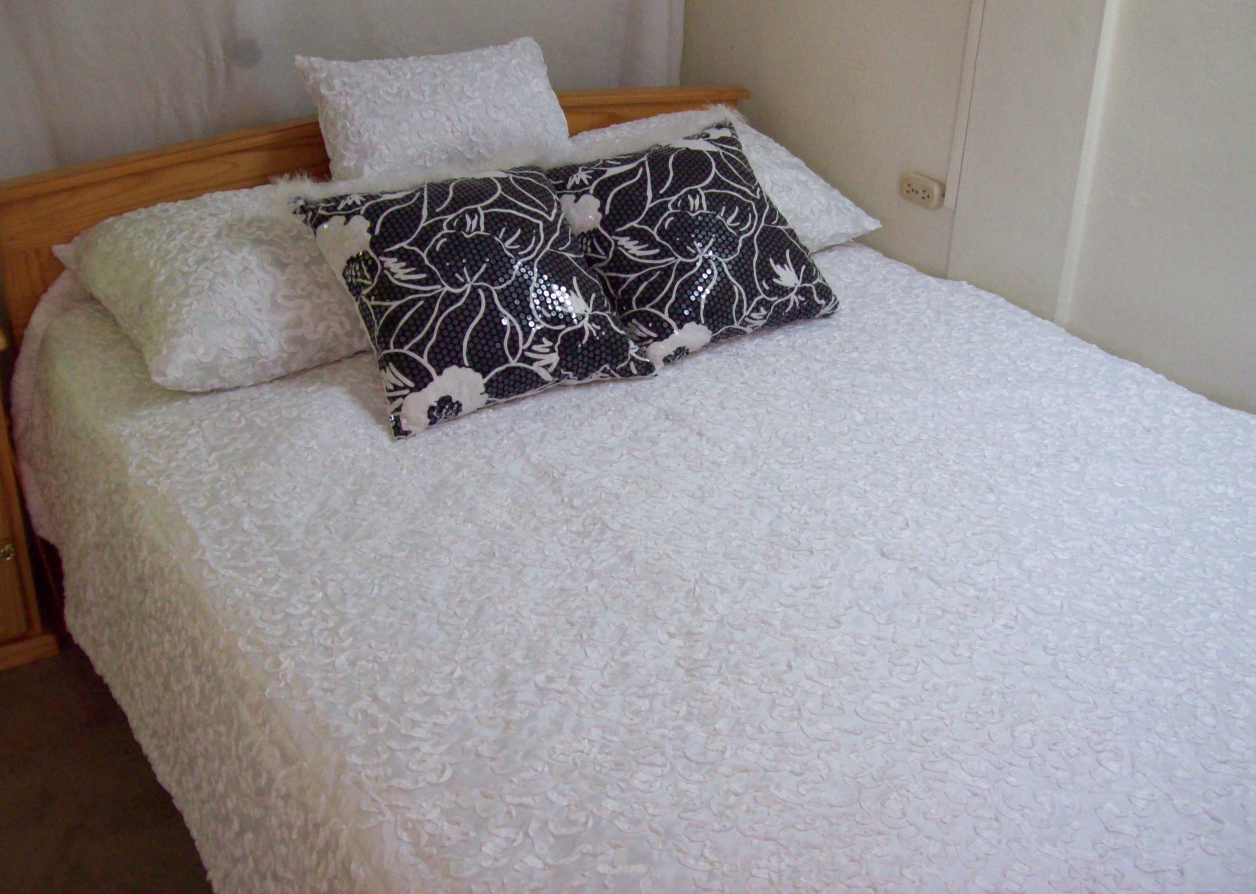 A bed with a white textured bedspread, a wooden headboard, two white pillows with textured covers, and two decorative black and white pillows with floral patterns. An electrical outlet is visible on the wall beside the bed.
