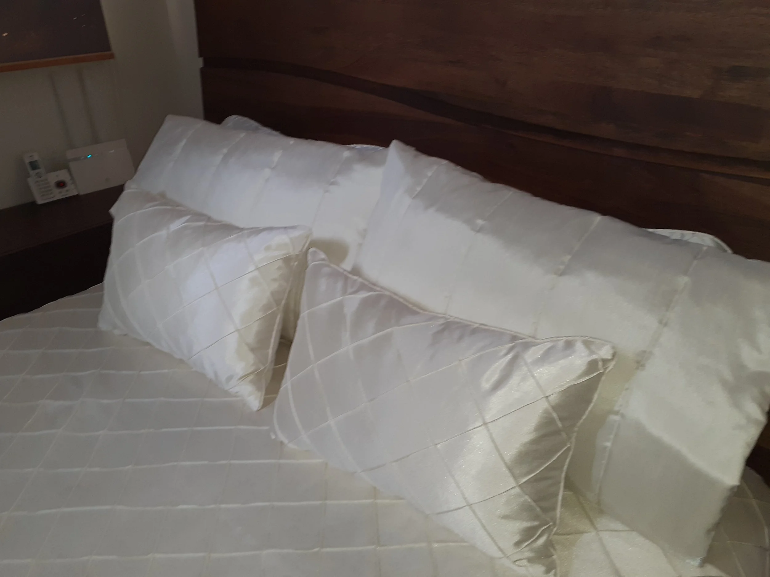 A bed with a white satin bedspread and matching pillows in a hotel room, with a dark wooden headboard.