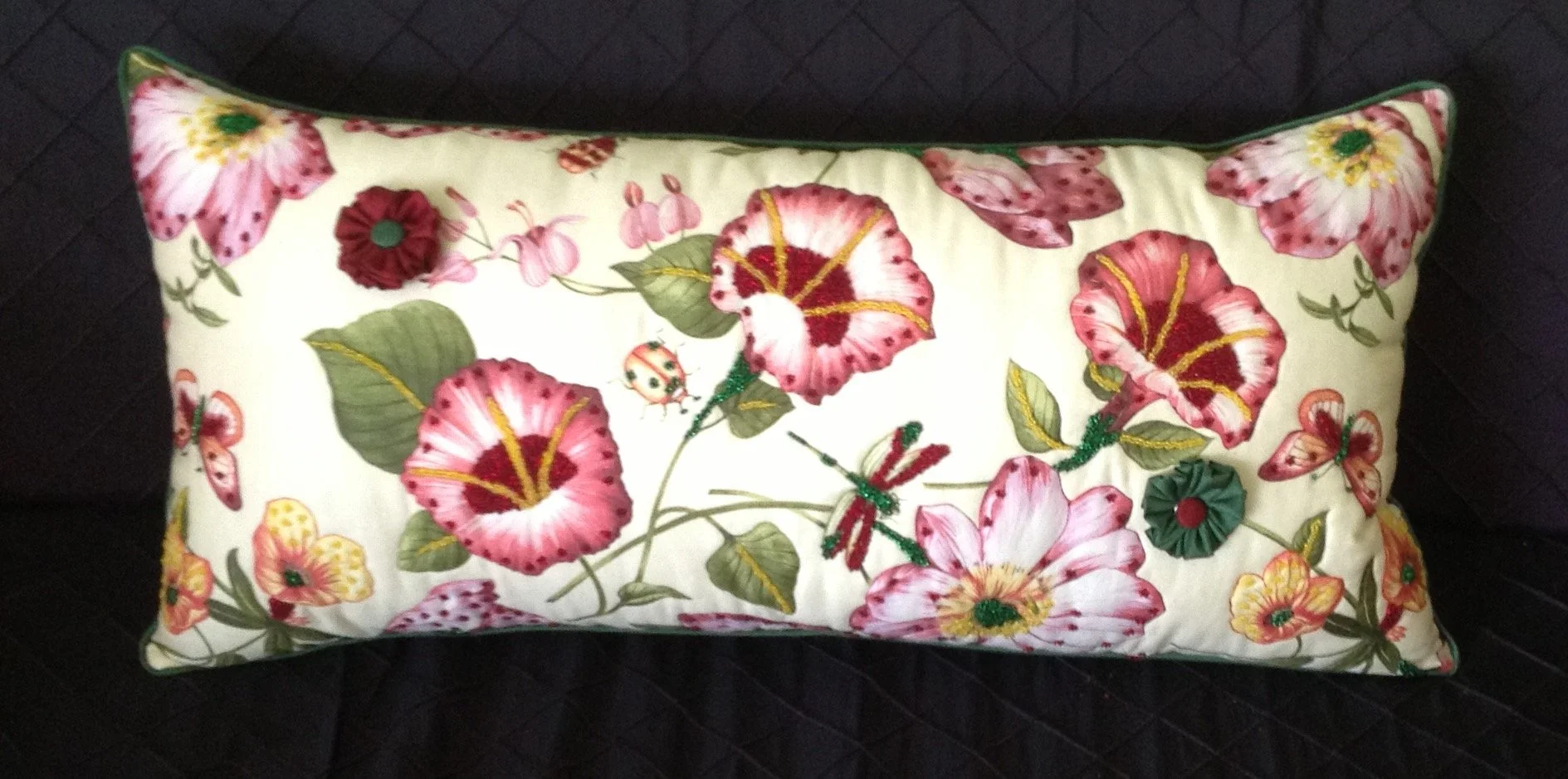 Decorative rectangular pillow with embroidered floral and butterfly design in pink, red, green, yellow, and white, resting on a black quilted surface.