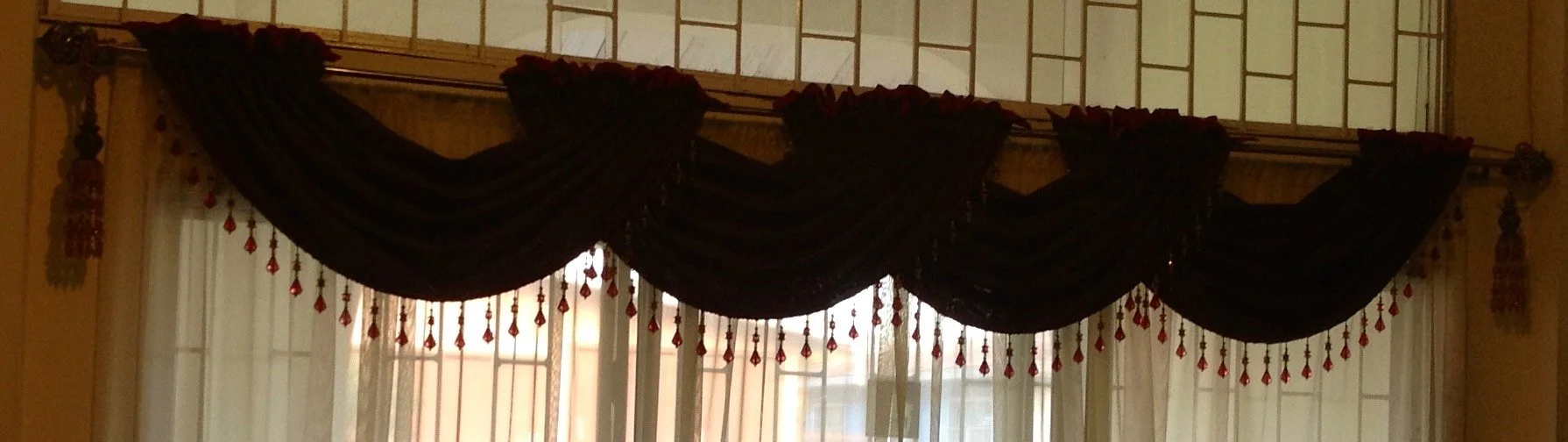 Black curtains with red tassels and beaded decorations, draped over a window with sheer curtains underneath.