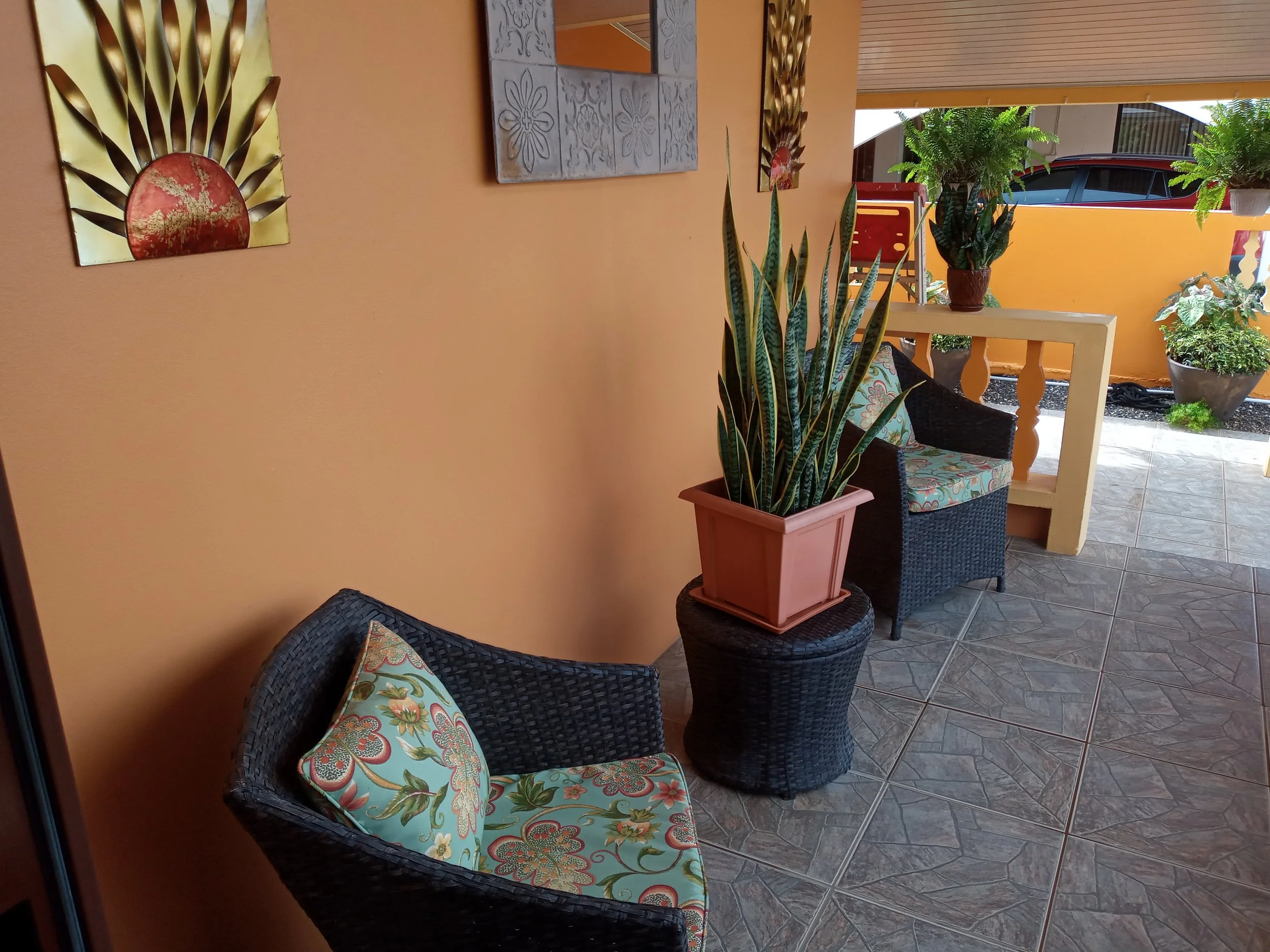 Decorative outdoor patio area with two black wicker chairs with floral cushions, a large potted snake plant, and potted ferns on a ledge, with yellow and orange walls, artwork, and a tiled floor.
