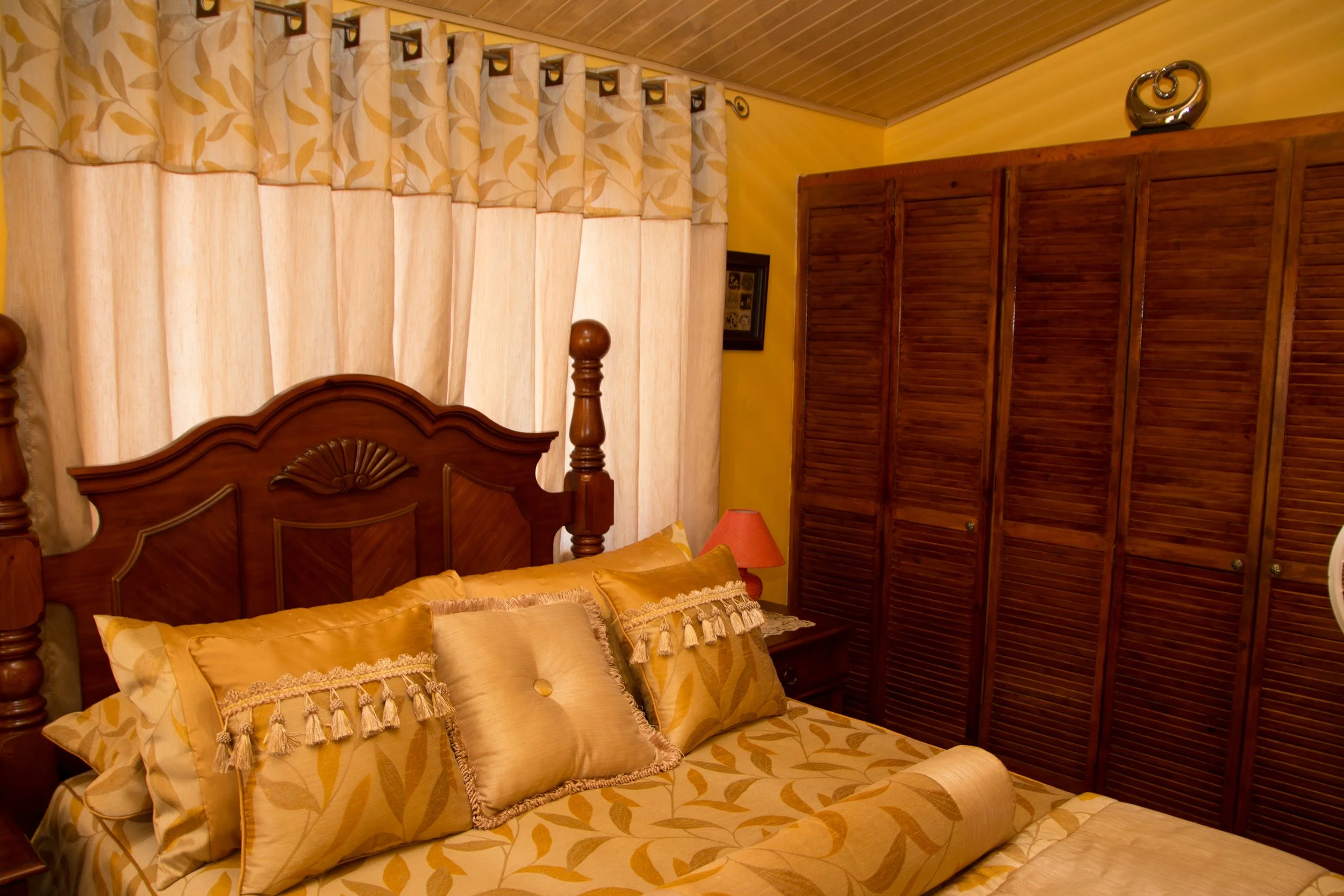 Interior view of a bedroom with a wooden bed, decorative pillows, a side table with an orange lamp, and wooden wardrobe doors.