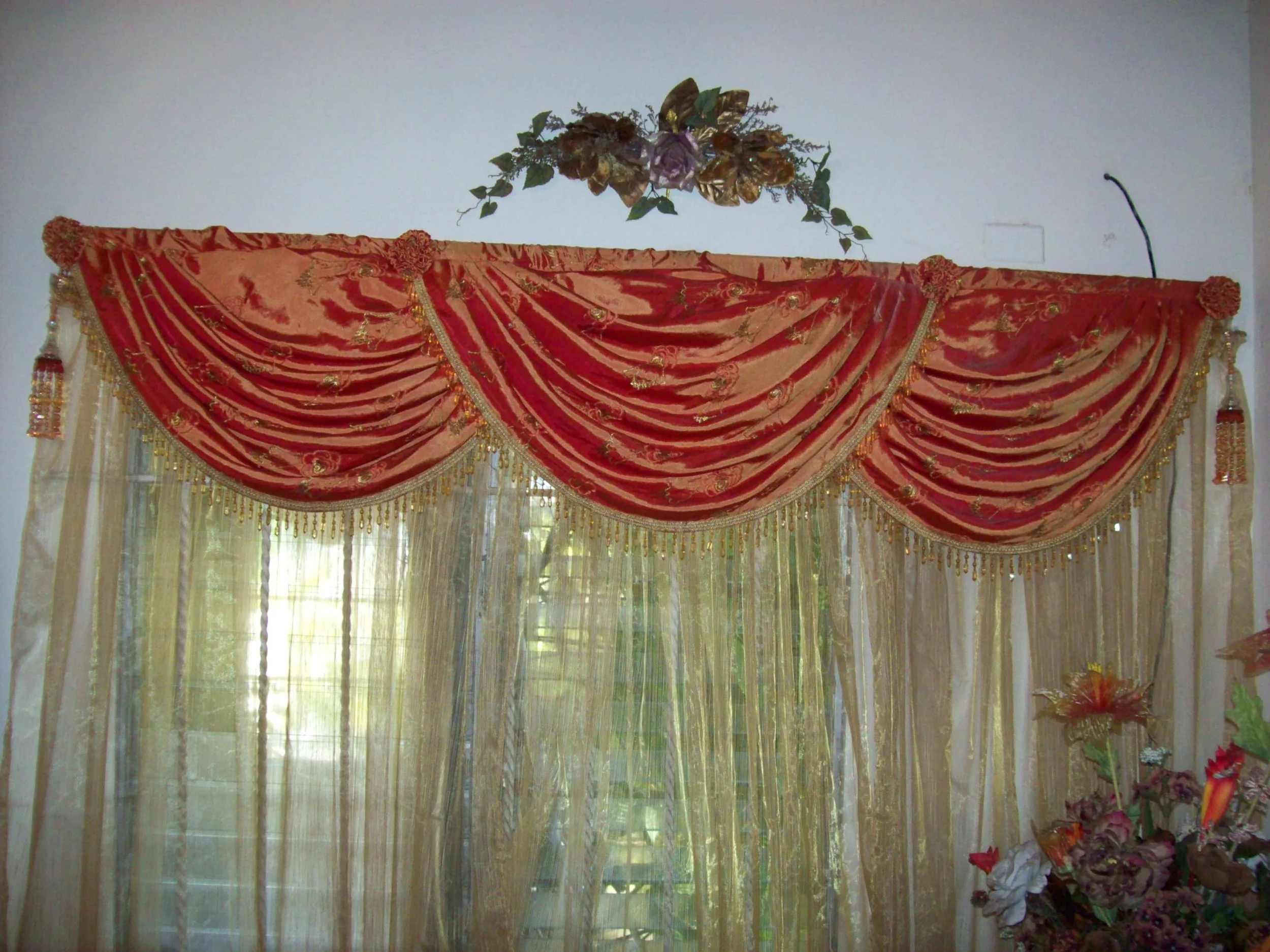 Decorative window curtain with red and gold drapes and sheer gold curtains, topped with a flower arrangement on the wall.