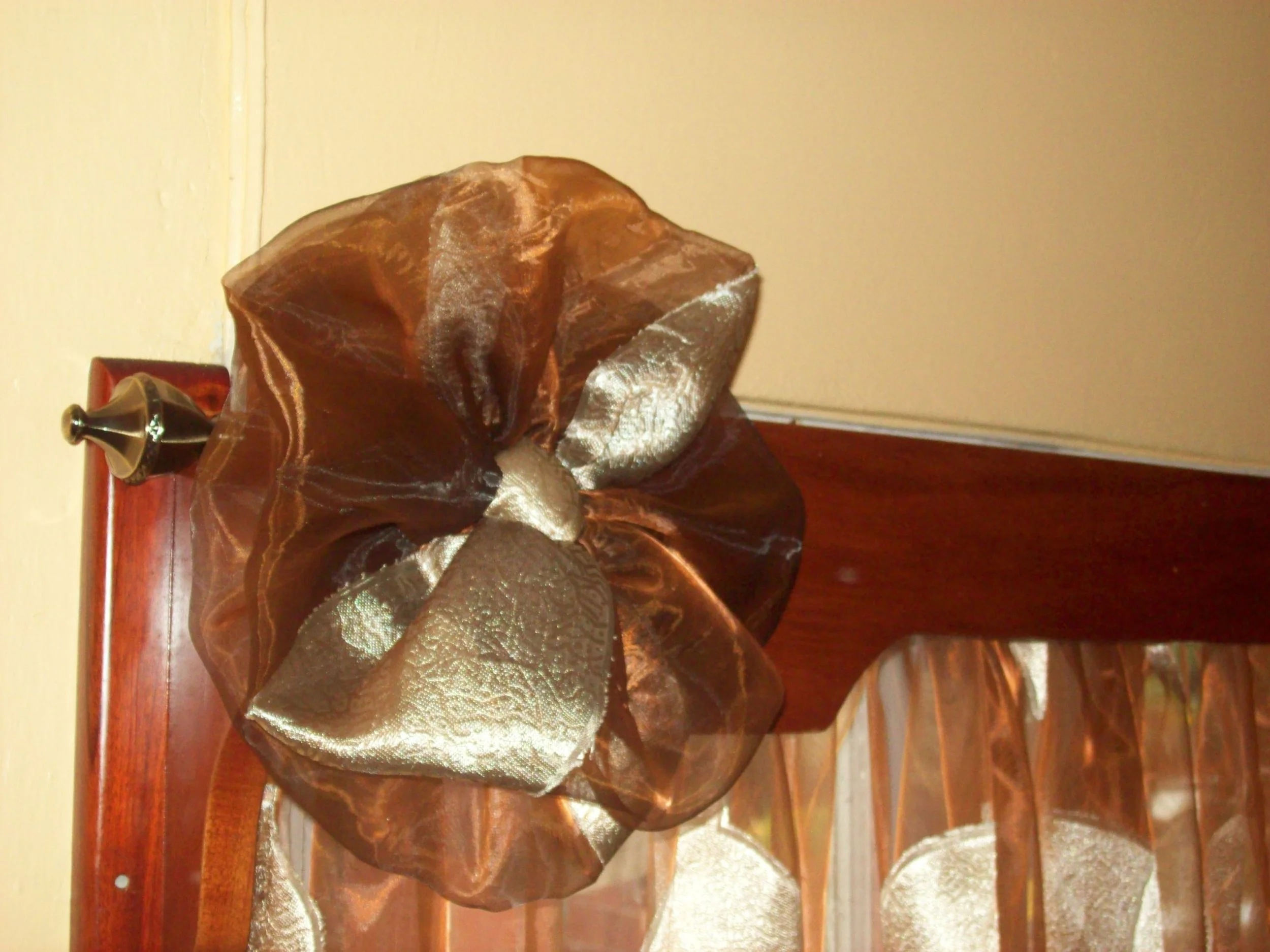Decorative sheer brown and gold curtain tieback with a metal hook, attached to a wooden window frame.