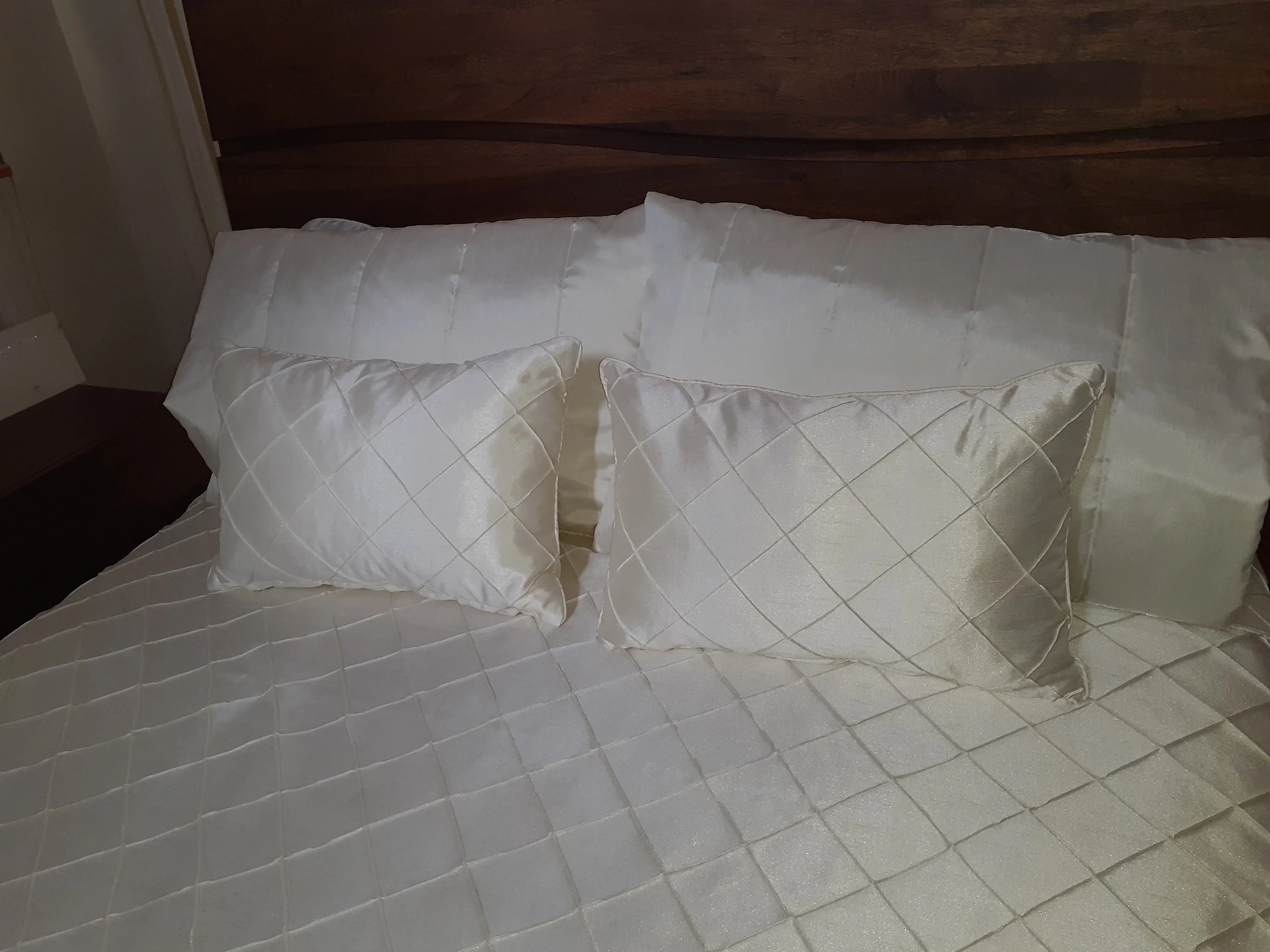 A close-up of a bed with white satin pillows and a matching satin bedspread with a diamond pattern, wooden headboard, and a wall in the background.