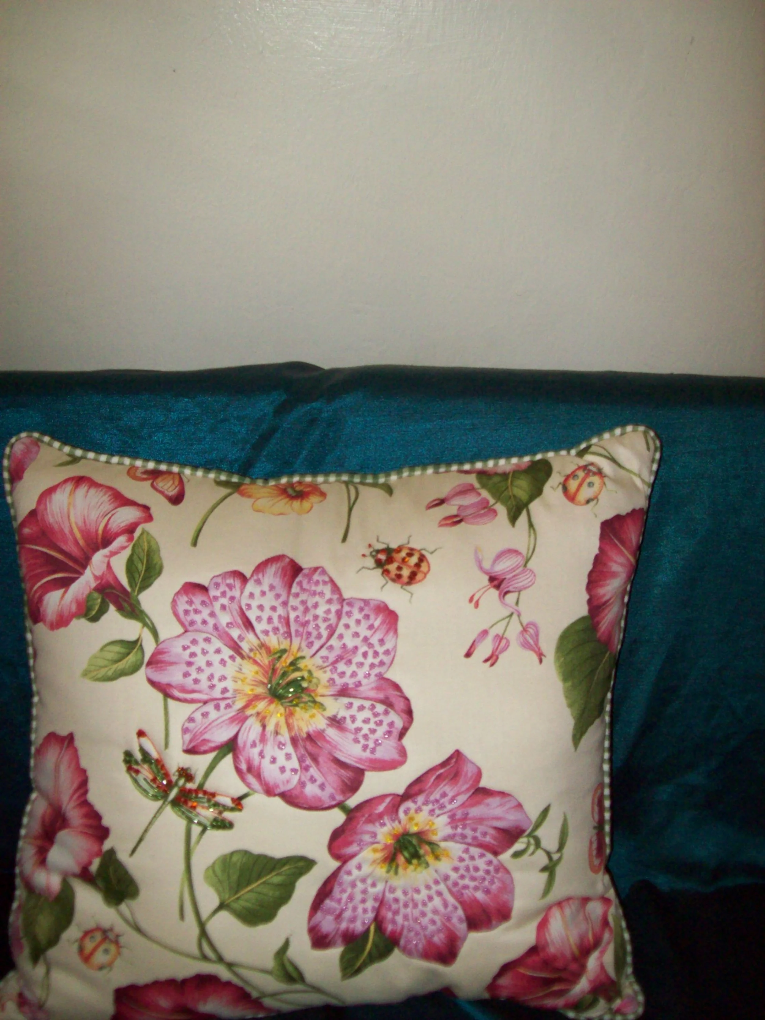 Decorative pillow with pink and purple flowers, ladybugs, and green leaves on a white background, resting on a teal sofa.