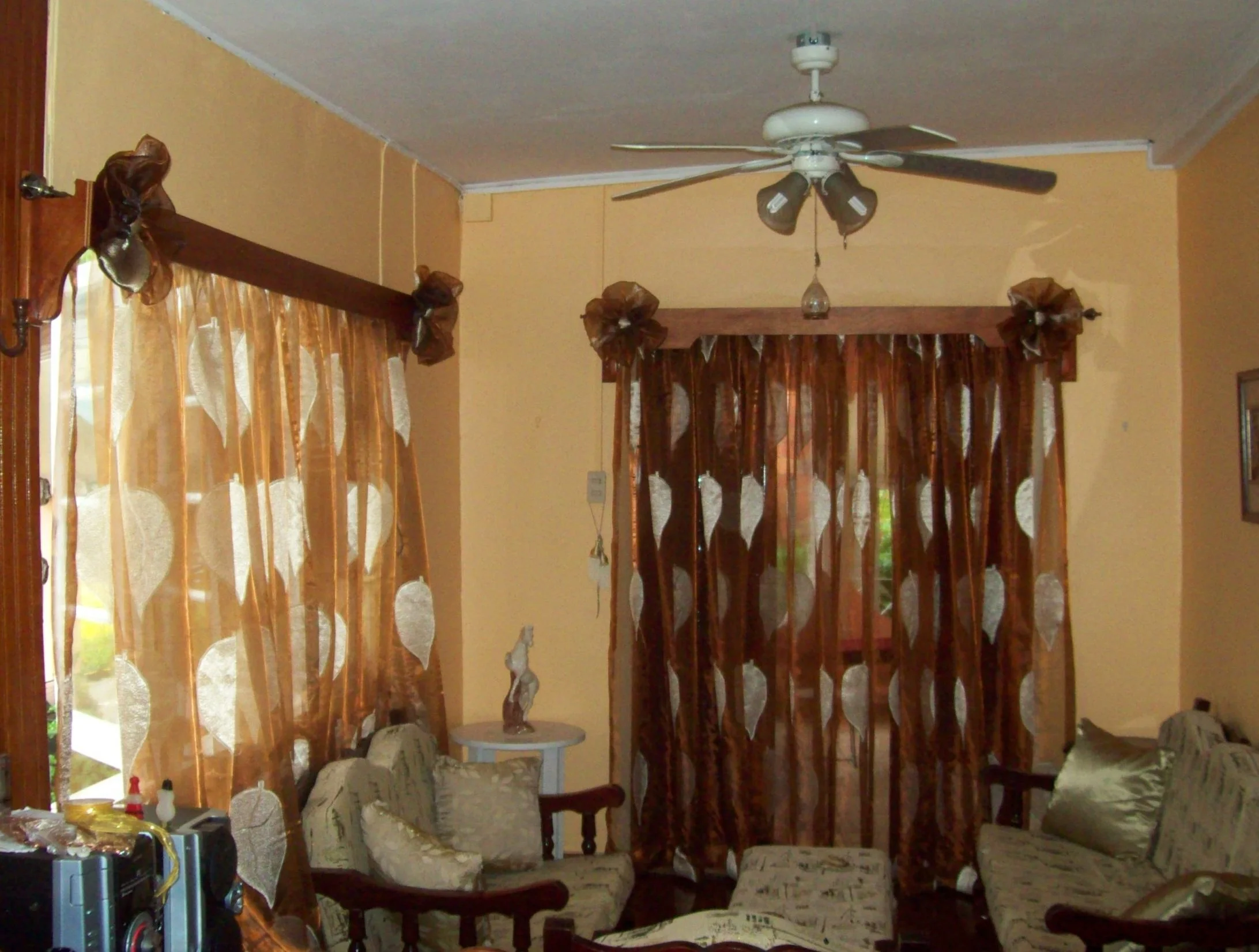 Living room with beige walls, brown curtains with leaf pattern, ceiling fan, sofa set, side table with statue, and some electronic devices.