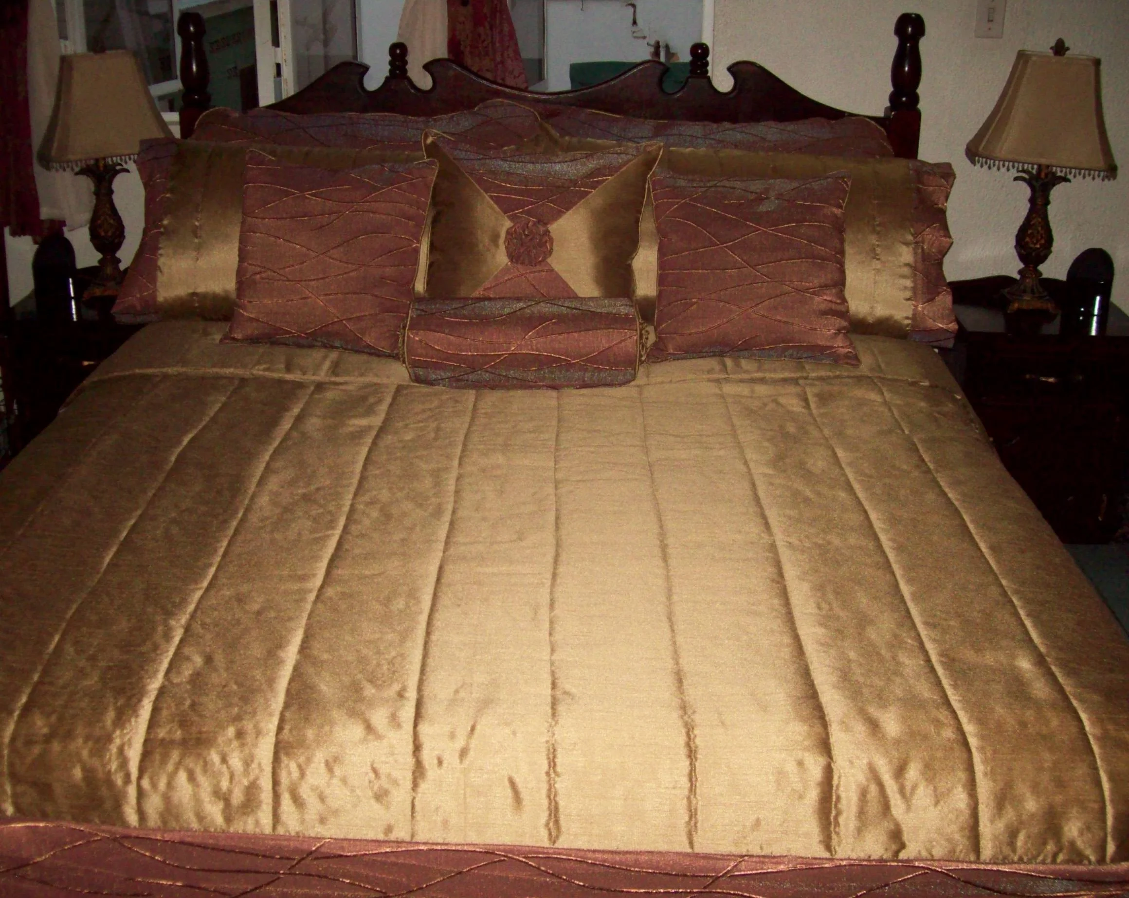 A bed with a brownish-gold quilt and multiple decorative pillows, with two matching lamps on nightstands on either side.