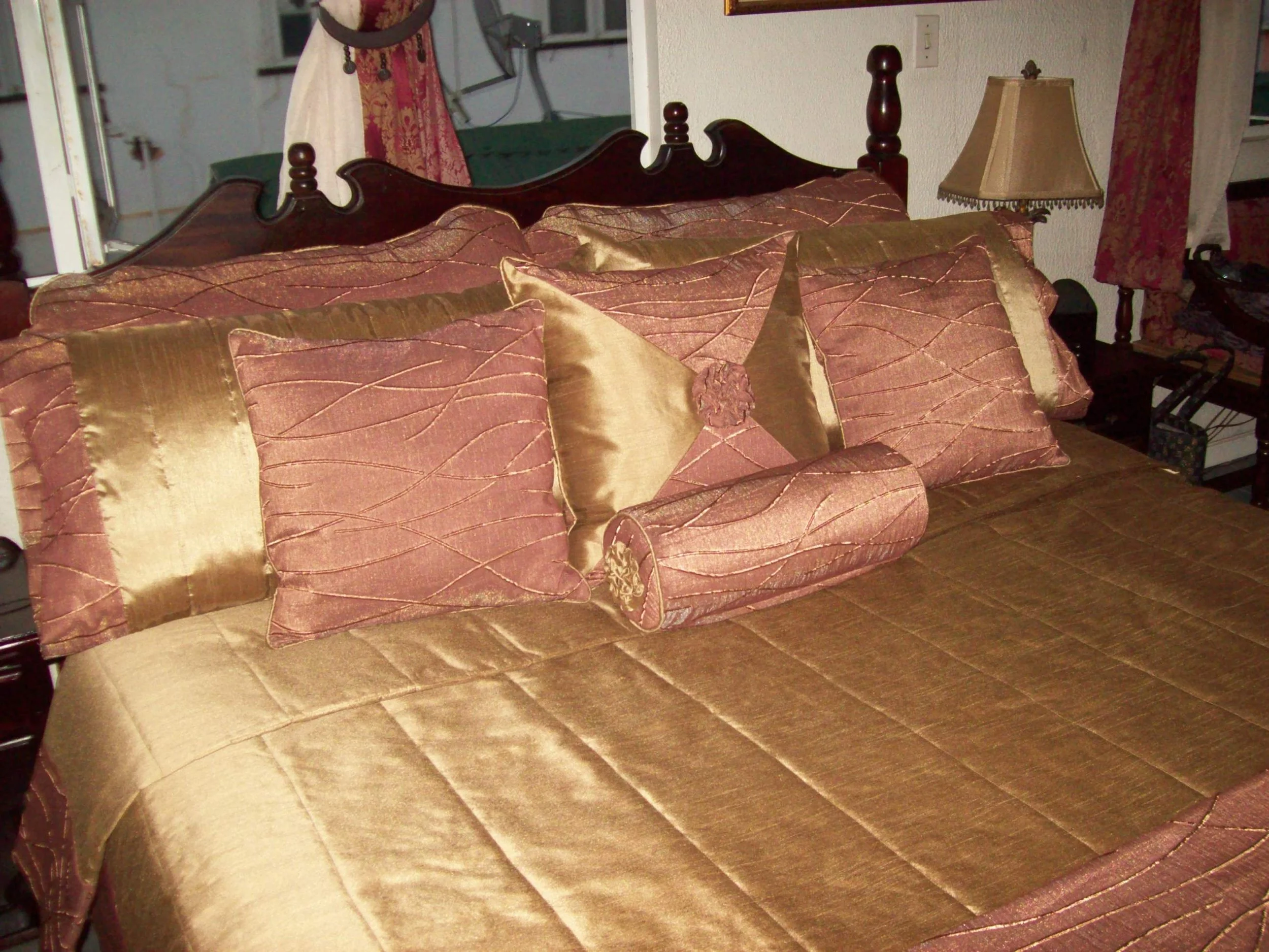 A bed with a wooden headboard, gold and maroon patterned bedding, and several decorative pillows, including cylindrical bolster pillows.