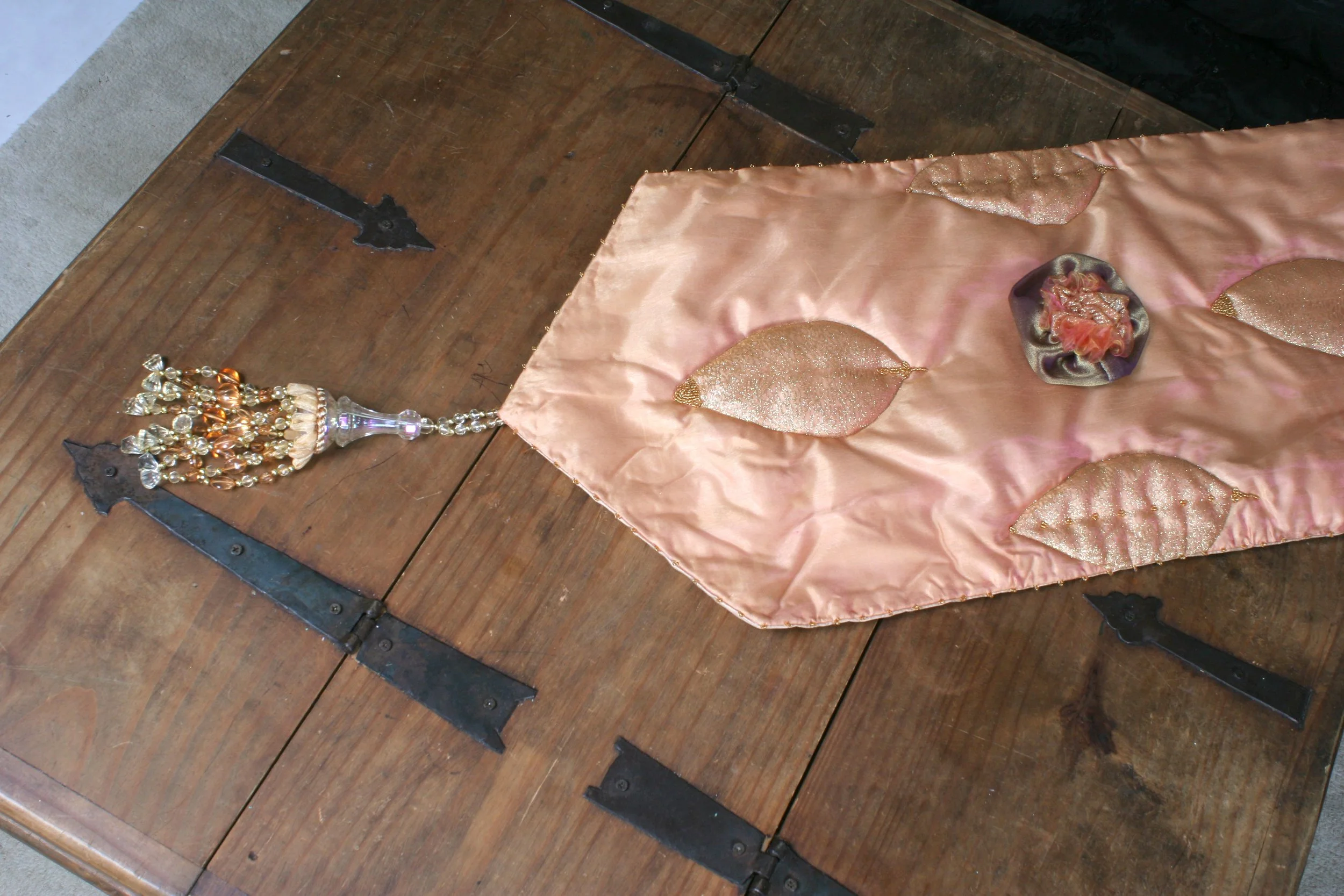 A wooden table with metal hinges has a pink satin fabric with gold appliqué leaf designs spread on it. An ornate beaded chandelier hangs from the fabric.