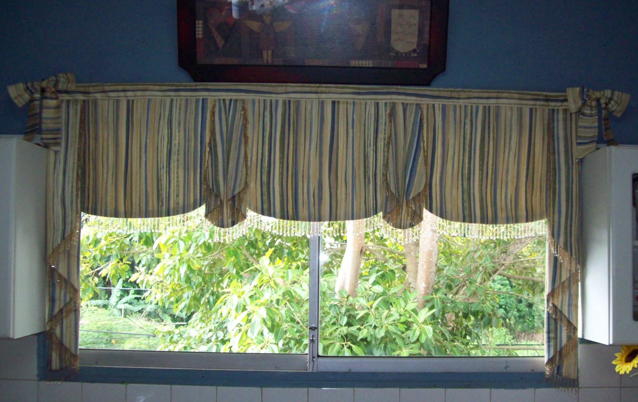 Kitchen window with striped valance curtain featuring blue, beige, and gold colors, tied in bows at the sides, and overlooking green foliage outside.