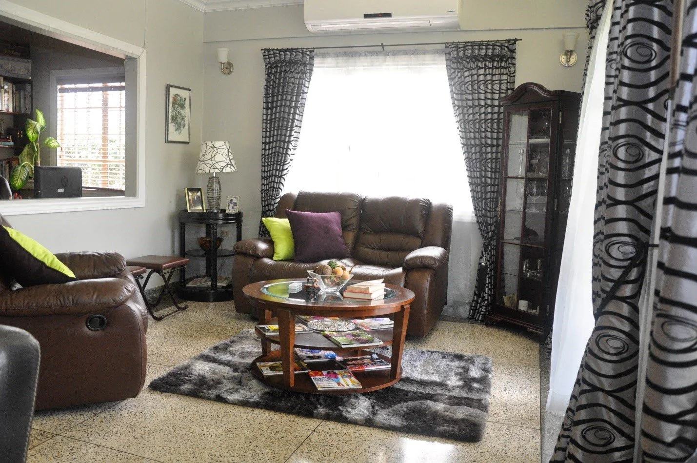Living room with brown leather sofa and armchair, black and white curtains, glass display cabinet, coffee table with books and decorations, positioned on a black and white rug, with a window letting in natural light.