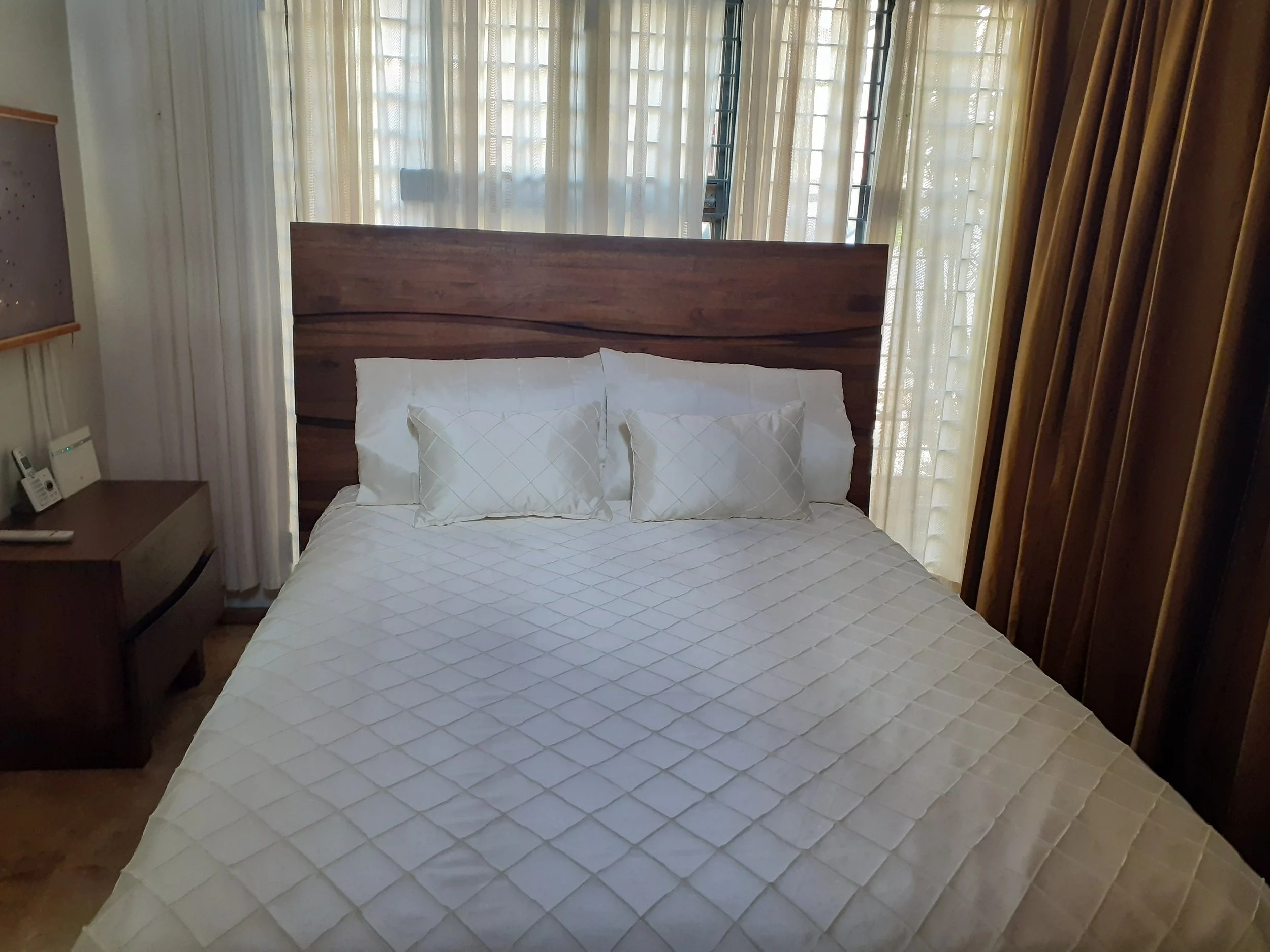 A neatly made bed with a wooden headboard, three white pillows, and a white quilt in a bedroom with light-colored curtains and a nightstand with a phone and a remote control.
