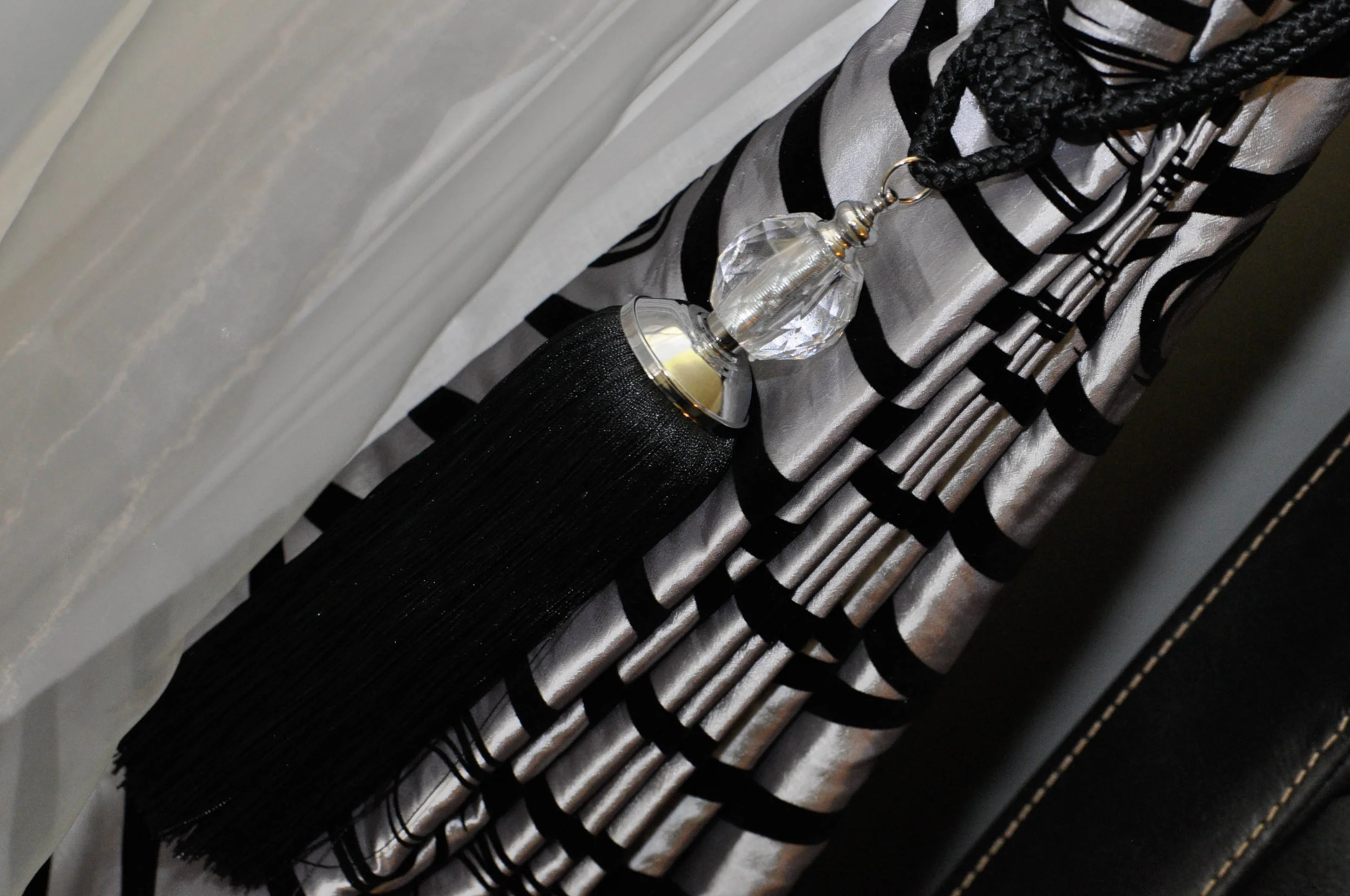 Close-up of a decorative curtain tieback with a crystal-like ornament and black fringed tassel, holding back a silver and black patterned curtain.