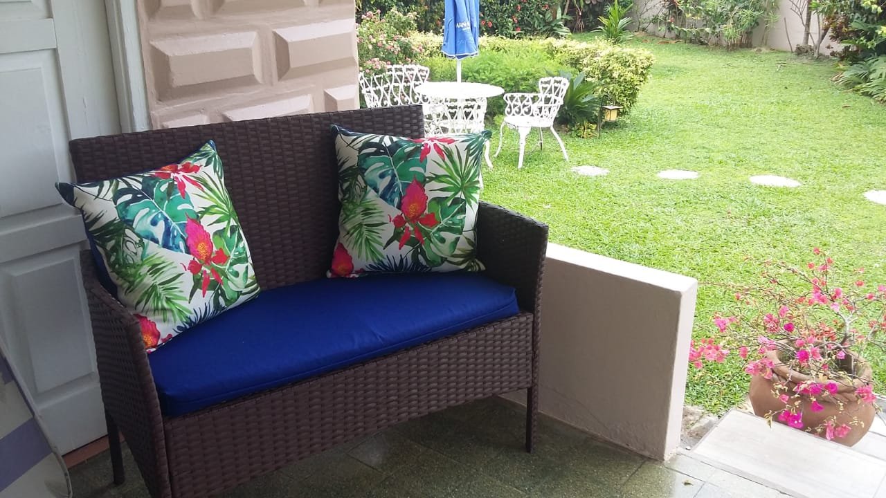 Patio area with a wicker bench with two tropical patterned pillows and a blue cushion, overlooking a green backyard with white outdoor furniture and a blue umbrella.