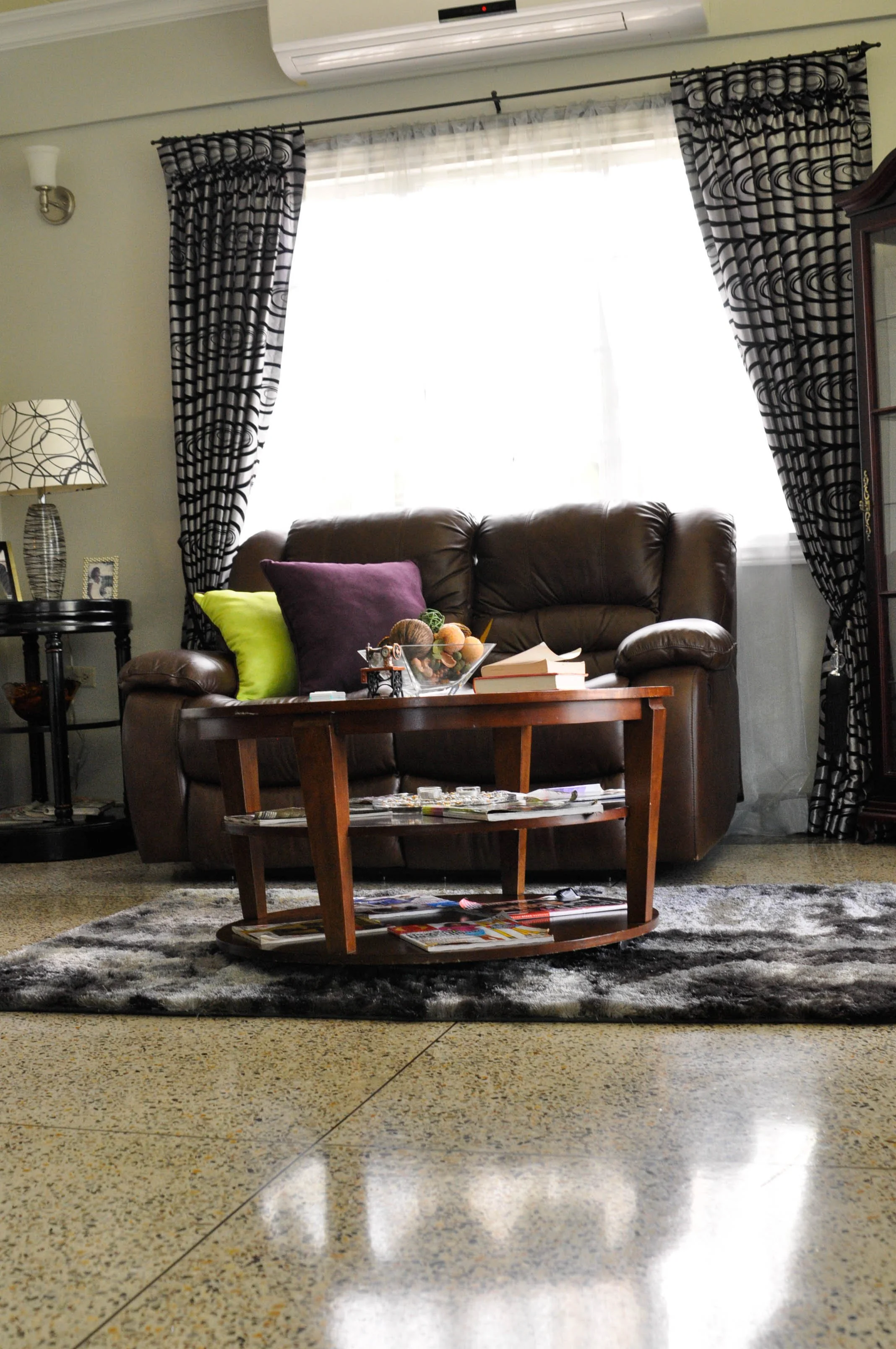 Living room with a brown leather sofa, purple and green cushions, a wooden coffee table with magazines and books, a patterned carpet, curtain and window, a side table with a lamp, and framed photos.