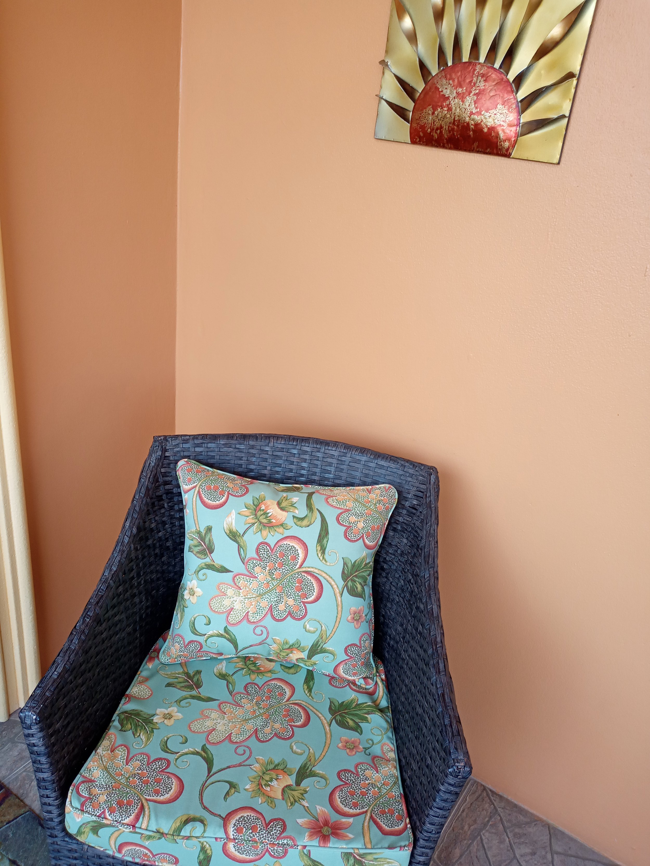 A wicker chair with a floral-patterned cushion and pillow, placed against a peach-colored wall. A metal wall art piece with a sun design is hung above the chair.
