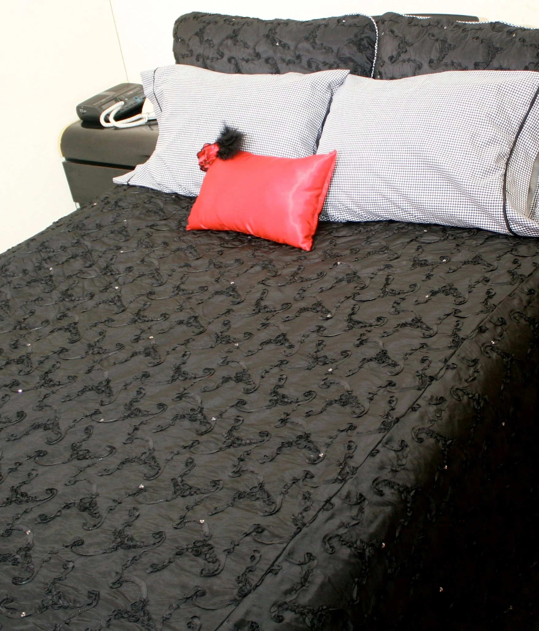 A bed with a black, embroidered quilt, two black-and-white checkered pillows, and a red satin pillow in front. A black hair extension with a red flower is on the pillows. There is a black nightstand with a telephone and a wall behind the bed.