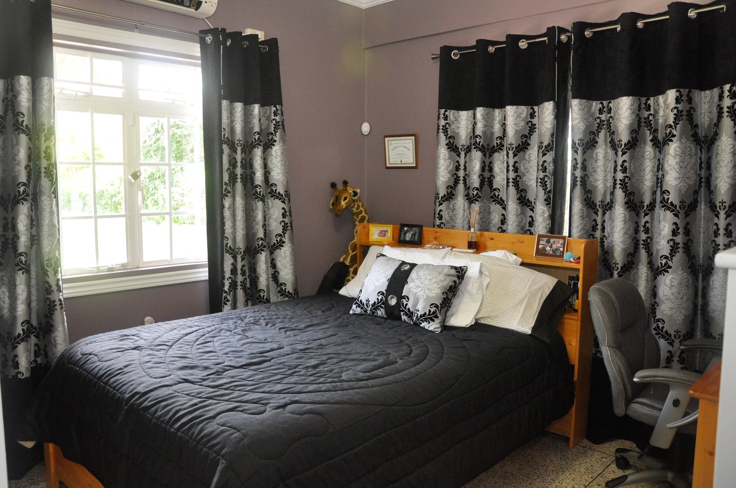 A bedroom with a bed, window with black and silver curtains, a stuffed giraffe, framed pictures, and a gray office chair.