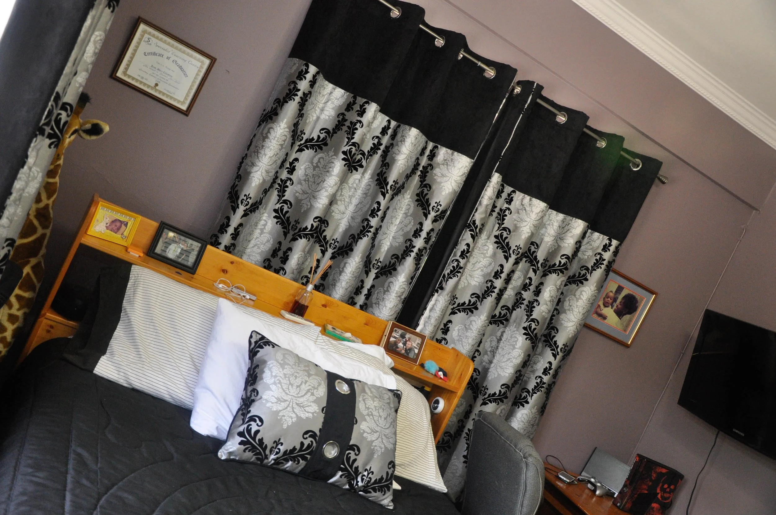 A bedroom with a bed, pillows, a wooden headboard with framed photos and personal items, curtains with black and white pattern, and wall art.