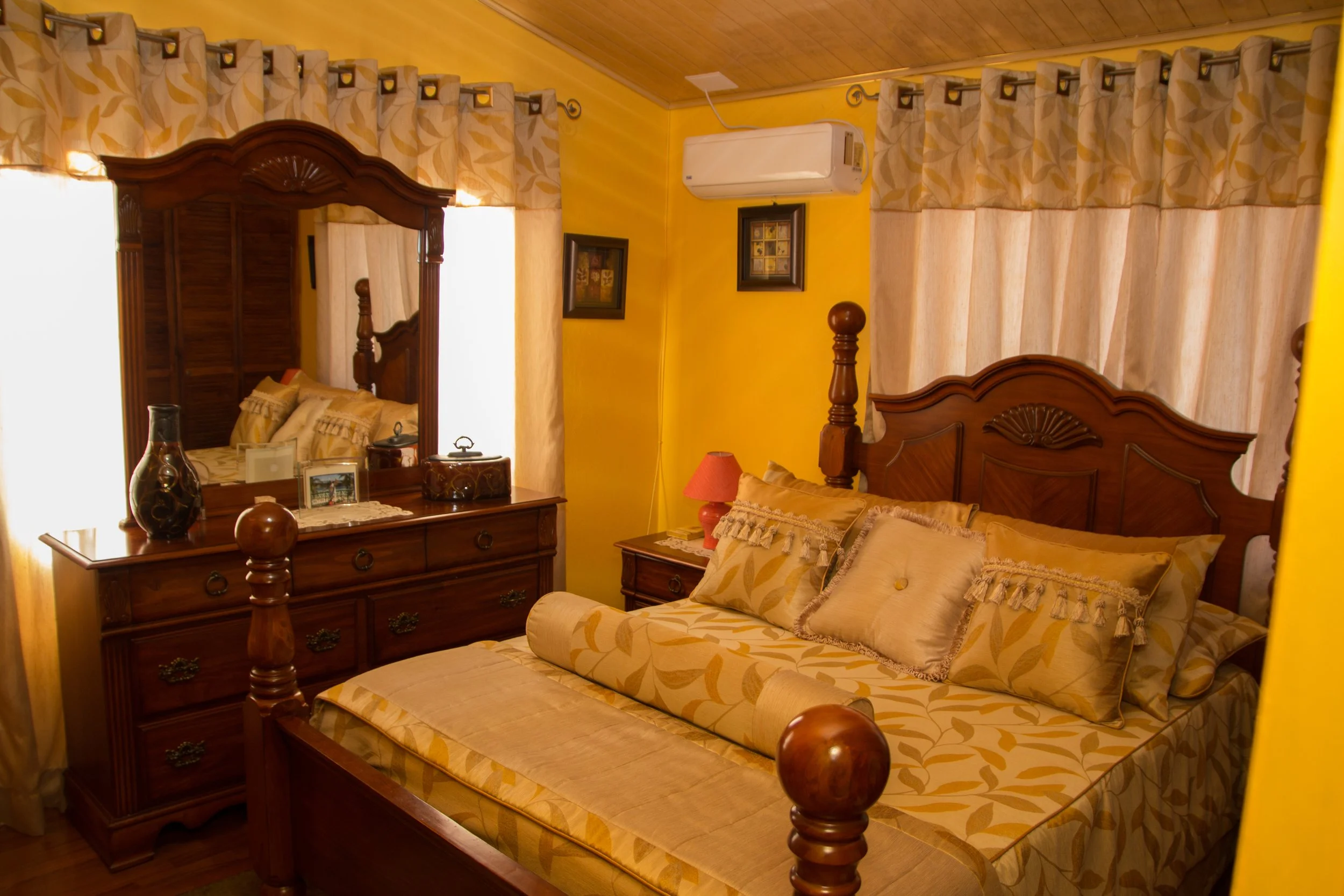 A bedroom with a wooden bed and matching dresser, yellow walls, curtains, a nightstand with a lamp, and framed pictures on the wall.