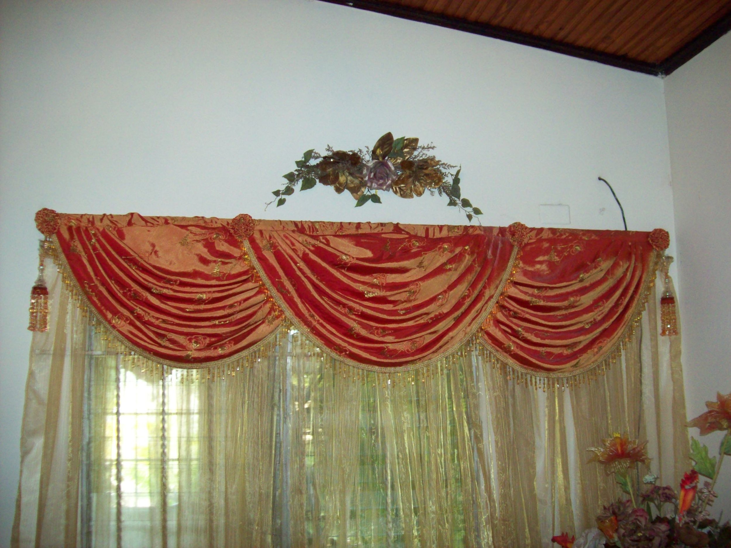 Decorative window curtain with red velvet drapes and gold embroidery, topped with floral valance in a room with white walls, wood ceiling, and a floral arrangement.