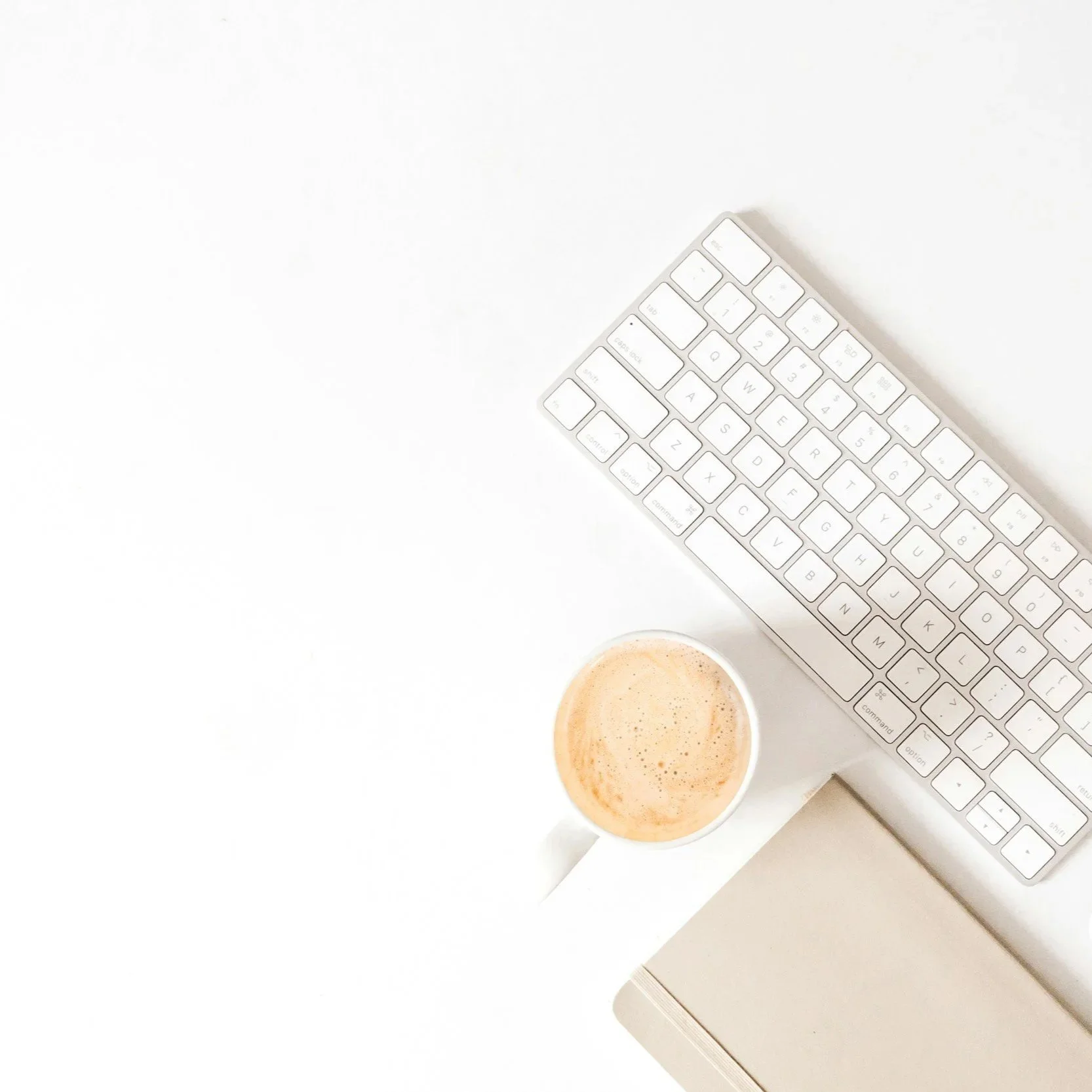 Overhead view of a white desk with a wireless keyboard, a cup of coffee, and a closed notebook.