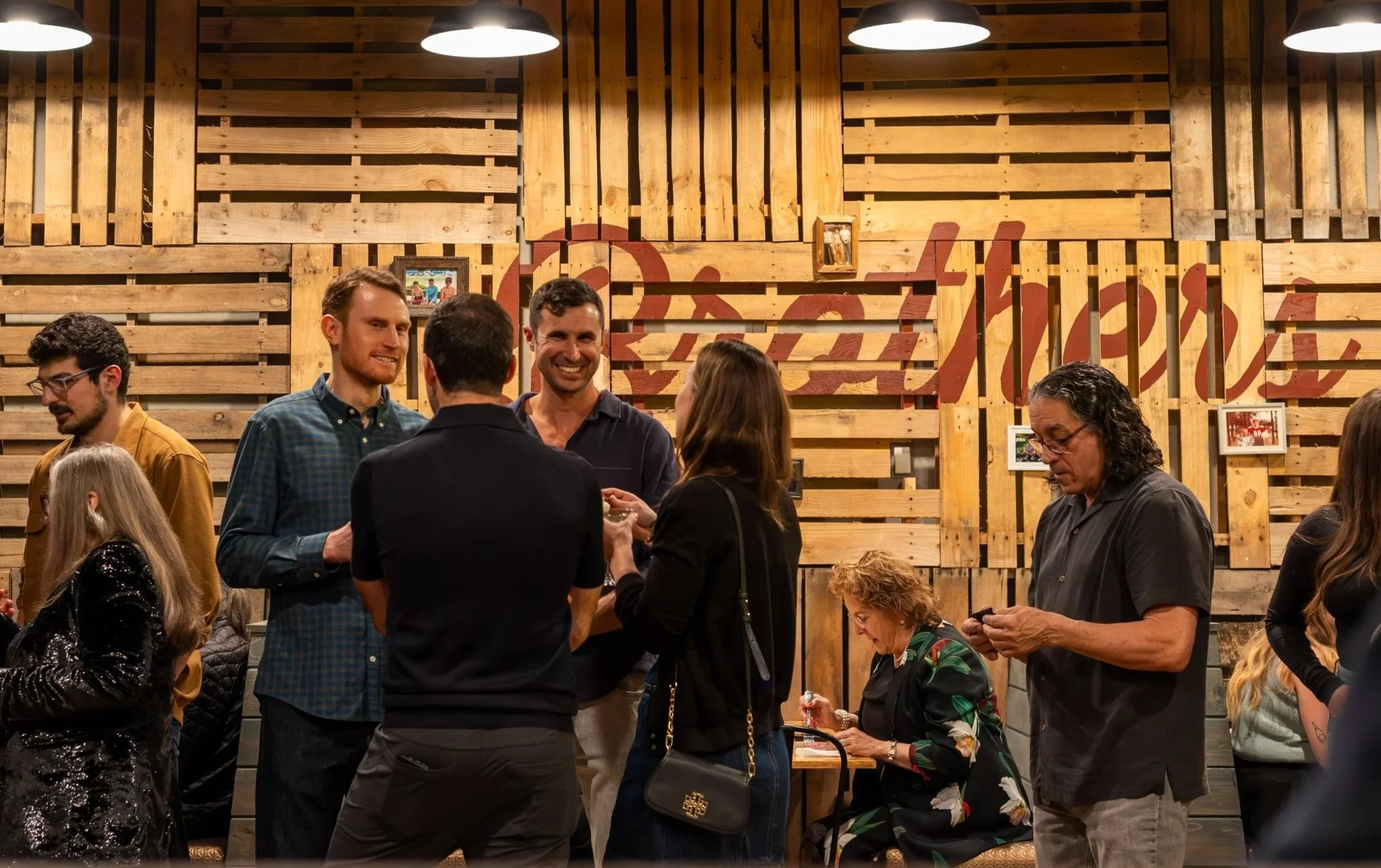 People socializing at a gathering in Brothers Meethouse with some engaged in conversation and eating Arkansas BBQ.