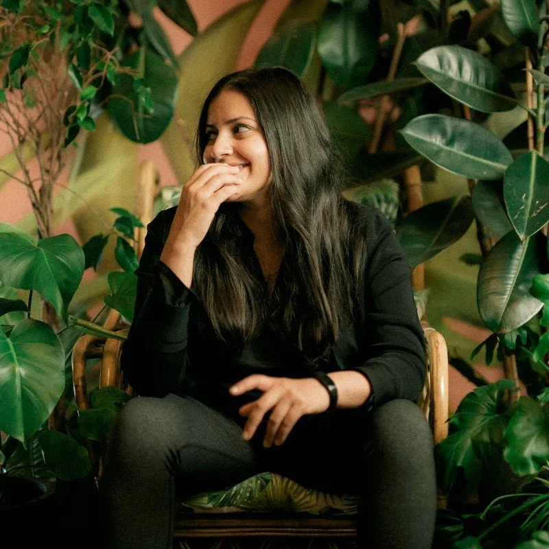 A woman with long dark hair smiling while sitting in a rattan chair surrounded by large green plants.