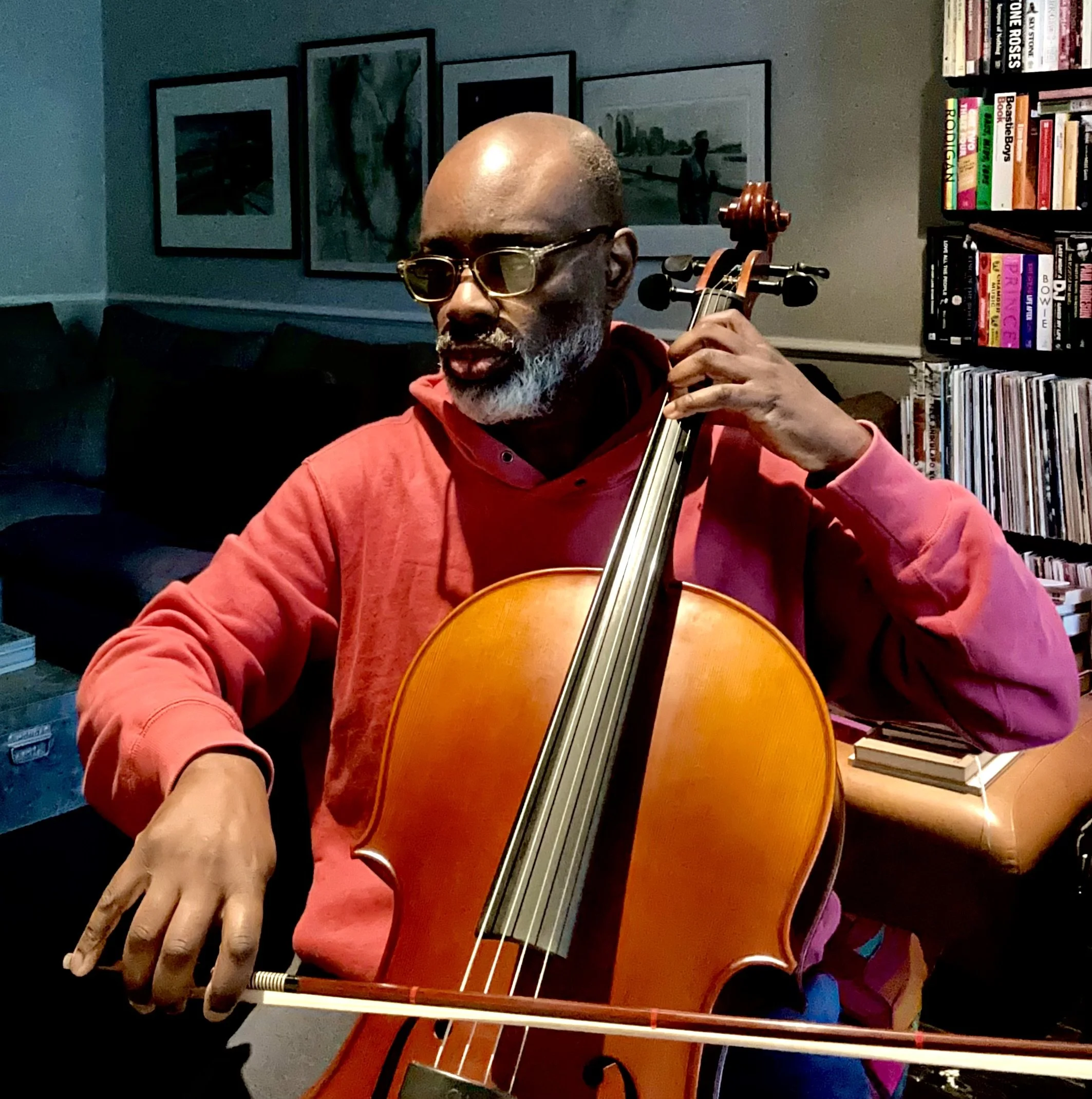 A man with glasses and a gray beard playing a cello in an indoor room with framed artwork on the wall and a bookshelf filled with books.