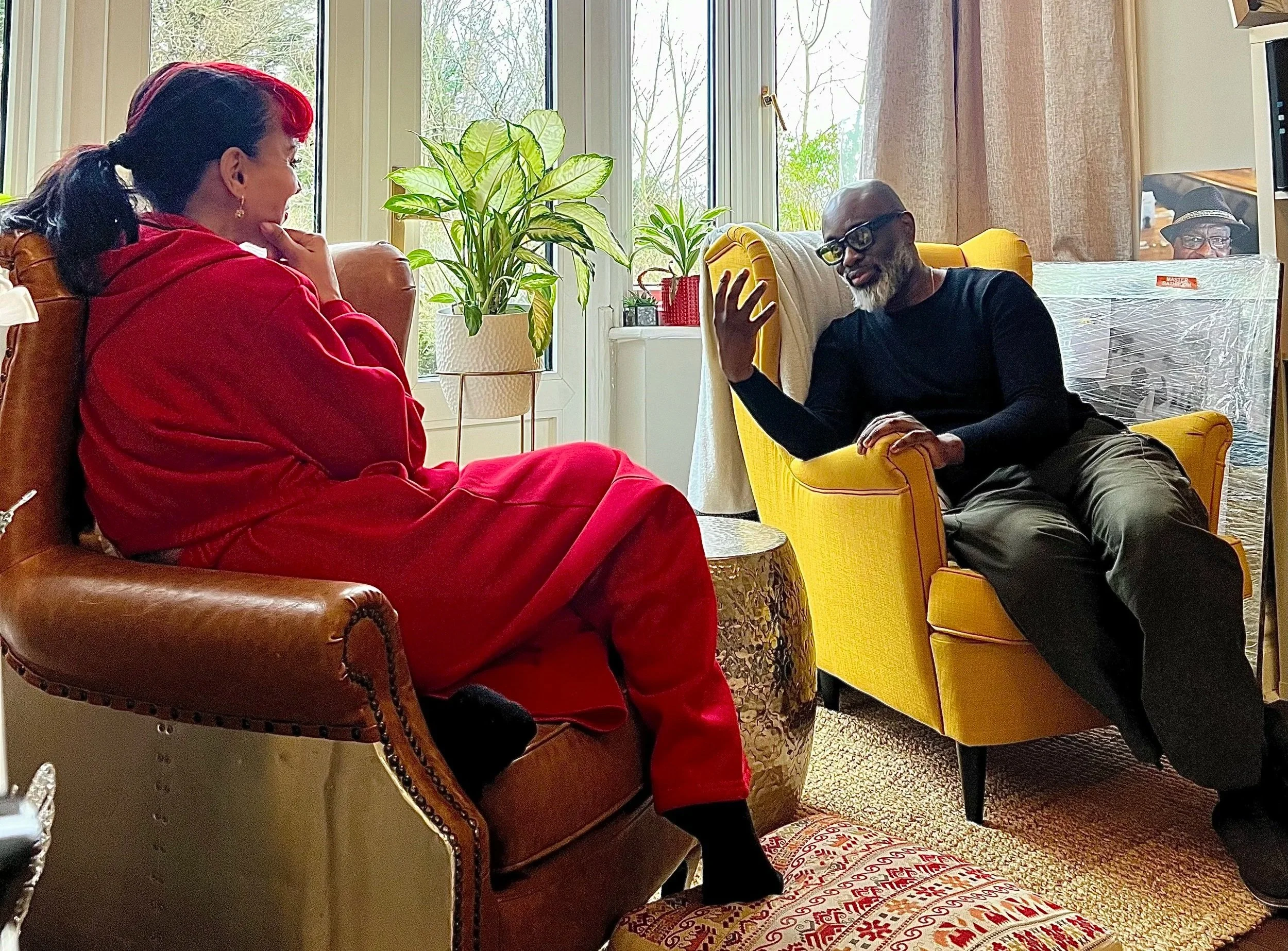 Two people having a conversation in a cozy living room, one woman in a red hoodie sitting on a brown leather armchair and a man in black glasses and a black shirt sitting on a yellow armchair, with potted plants and a large window in the background.