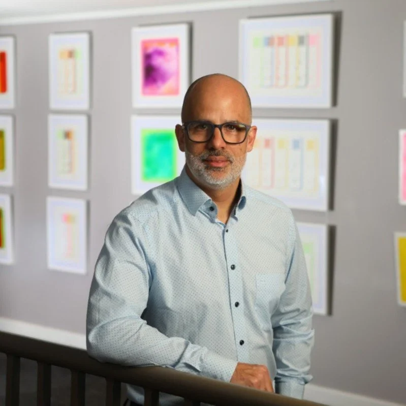 A middle-aged man with glasses and a gray beard stands in front of a wall with framed colorful abstract artwork.