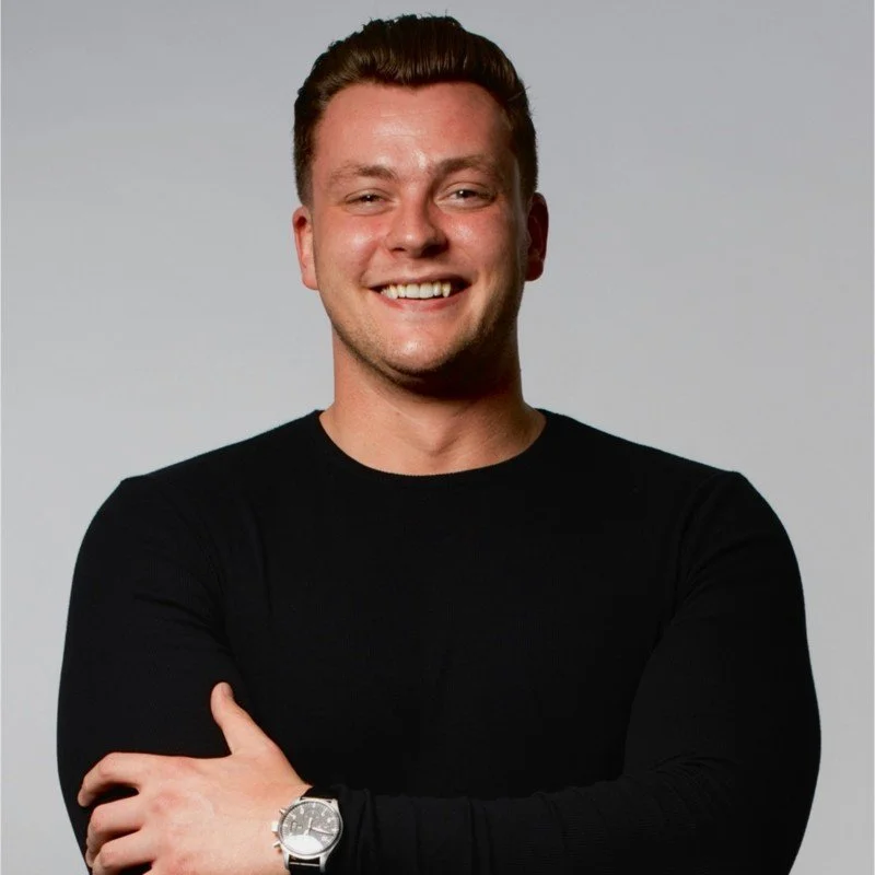 A smiling young man with short dark hair, wearing a black long-sleeve shirt and a silver watch on his left wrist, standing with arms crossed against a plain light gray background.