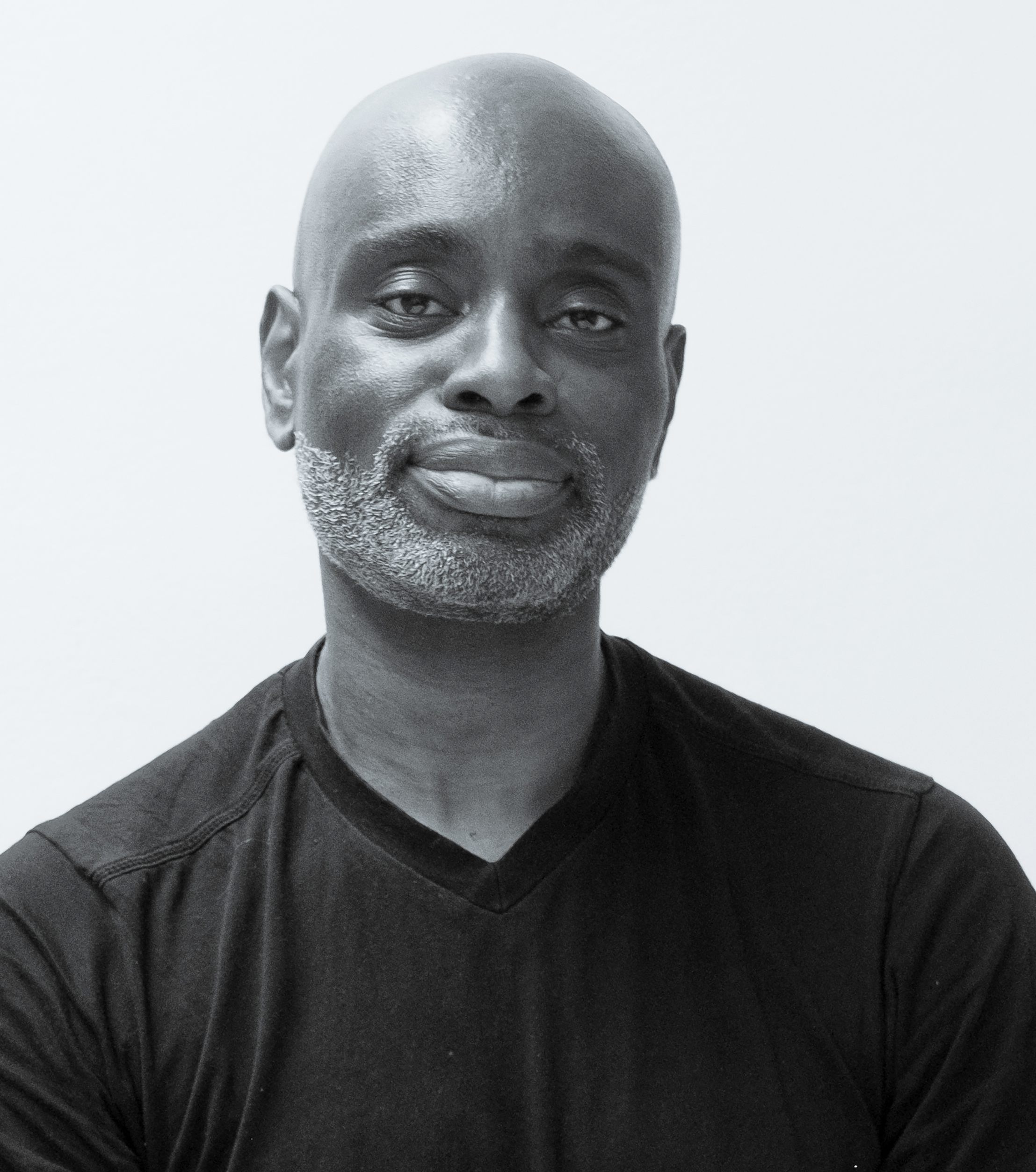A Black man with a short beard and bald head, wearing a black V-neck shirt, looking at the camera with a neutral expression against a plain light background.