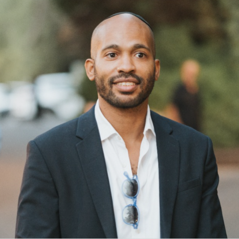 A man with a shaved head and beard wearing a black blazer, white shirt, and sunglasses hanging from his shirt, smiling outdoors.
