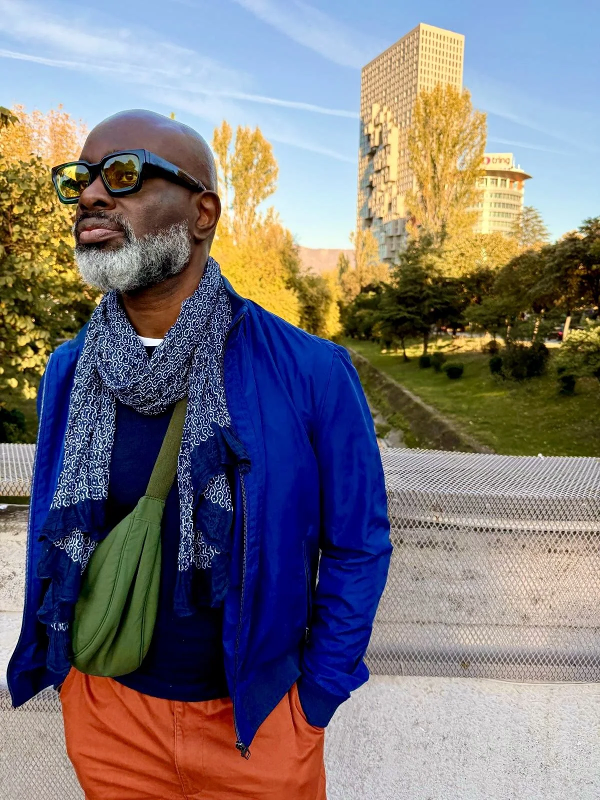 An older man with a beard and glasses standing outdoors on a bridge during daytime, wearing a blue jacket, scarf, and orange pants, with trees and a tall building in the background.