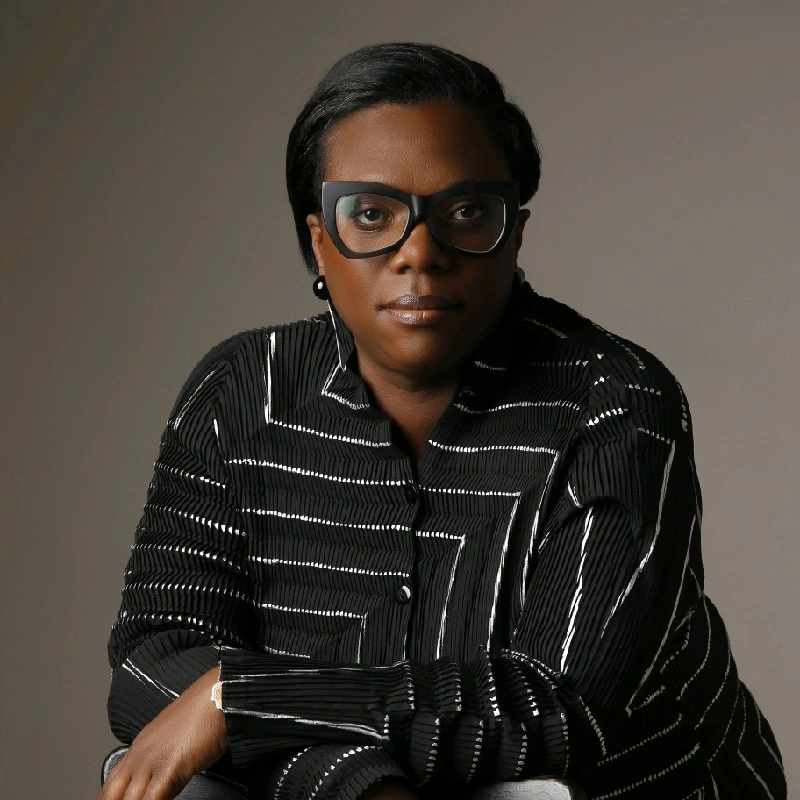 A woman with short black hair, wearing large black glasses and a black and white striped shirt, sitting with her arms crossed against a neutral background.