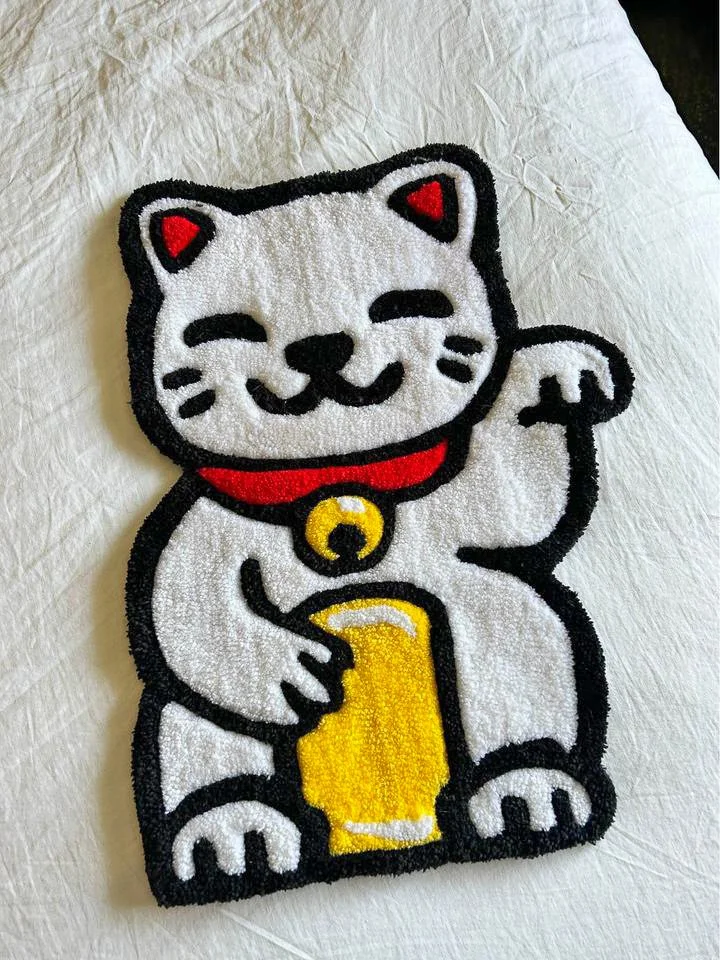 An embroidered patch of a smiling maneki-neko, a Japanese lucky cat, holding a beer mug, with a red collar and a yellow bell.