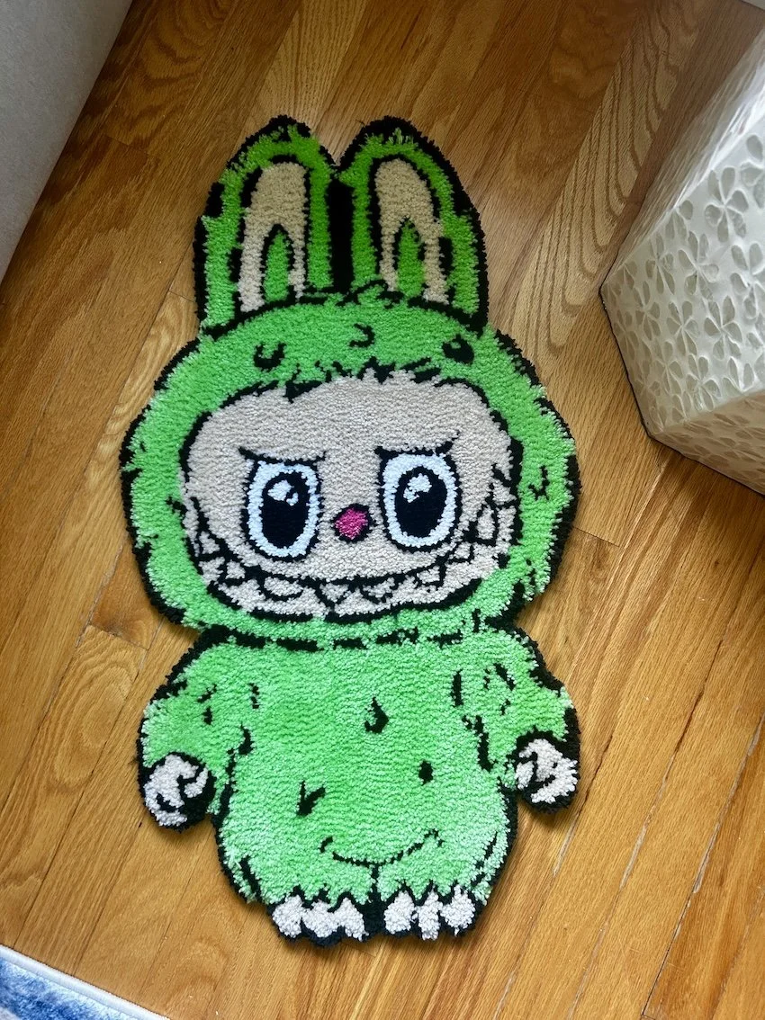 Colorful bath mat designed as a cute cartoon creature with large eyes, a pink nose, and green furry costume with hood and ears, on a wooden floor.