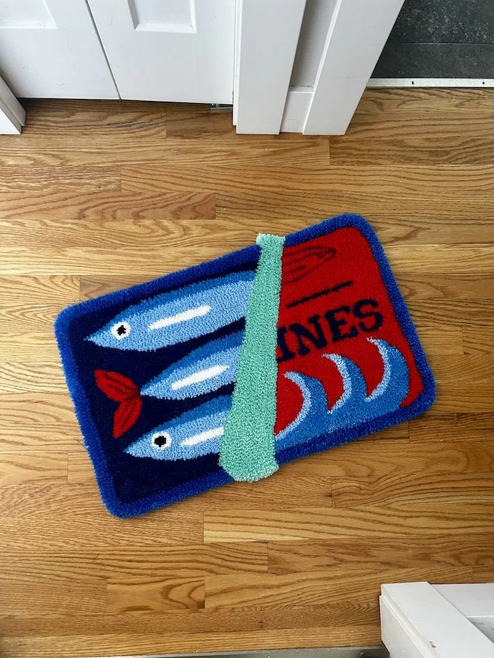 Colorful fish-shaped doormat with sea theme, featuring two fish and red claws, placed on a wooden floor near a door.