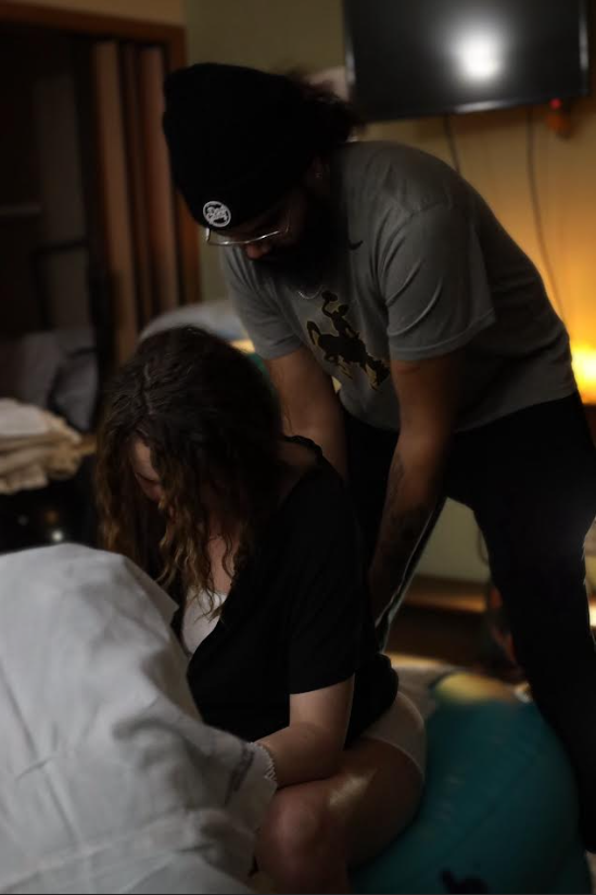 A person with curly hair sitting on a bed with their head down, another person standing next to them leaning over, wearing glasses, a beanie, and a gray t-shirt, in a dimly lit room.
