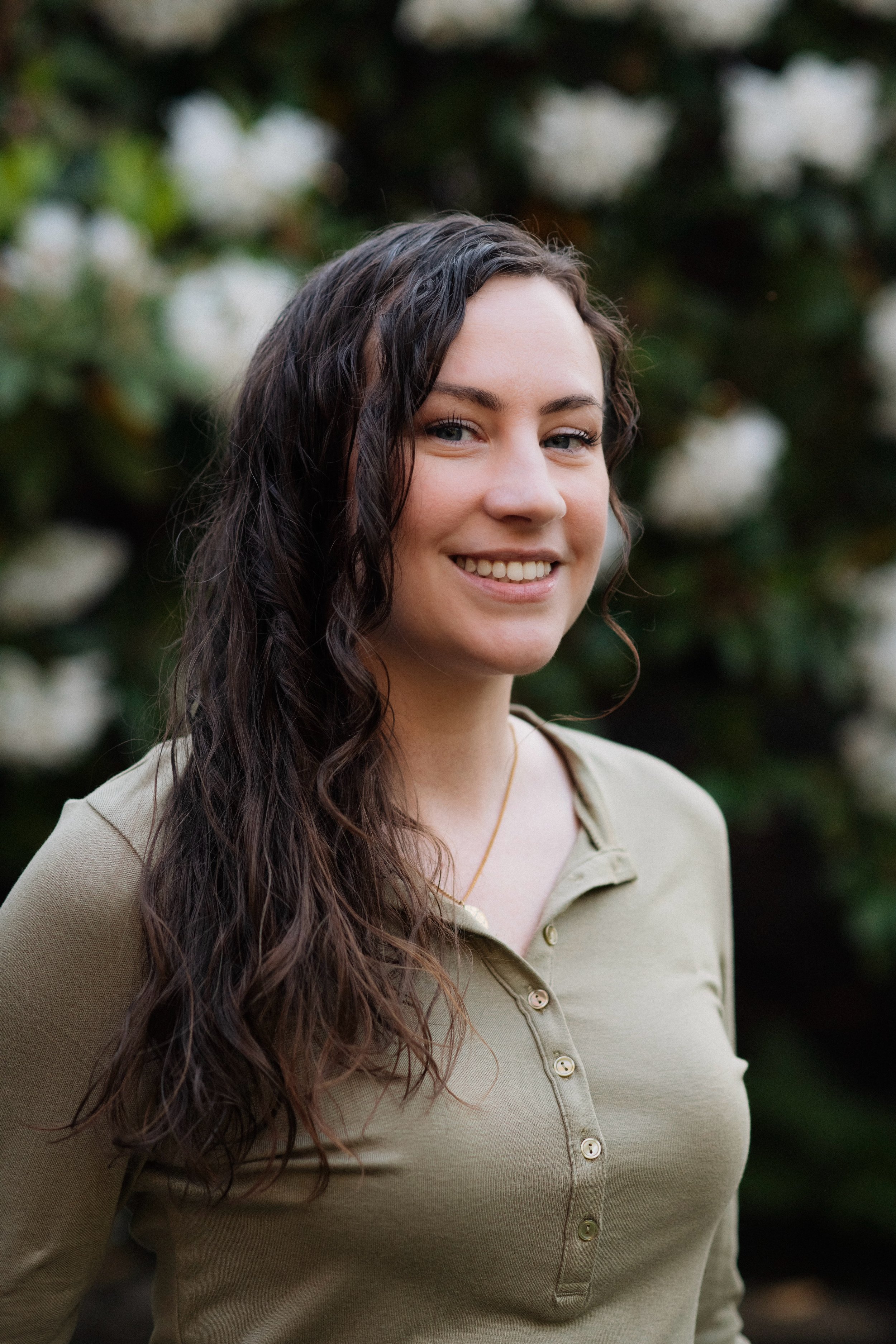 A young woman with long, curly brown hair smiling outdoors in front of blurred white flowers and green foliage.