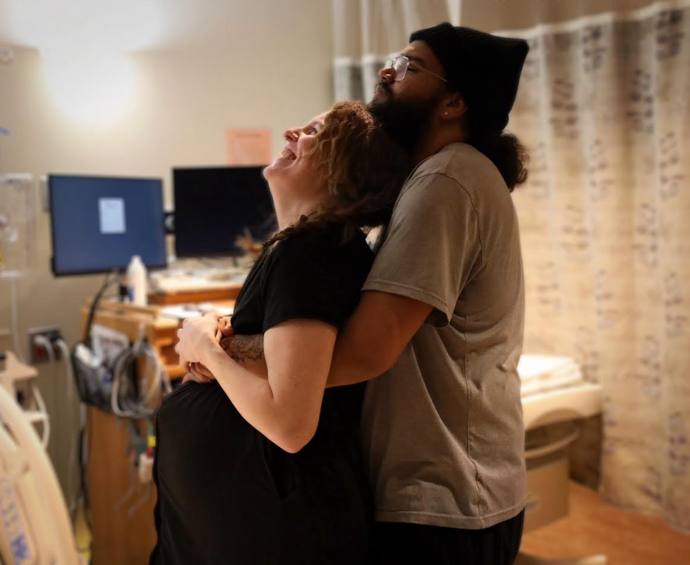 A couple, a woman and a man, happily embracing in a hospital room. The woman is pregnant, and they are smiling and sharing a joyful moment.