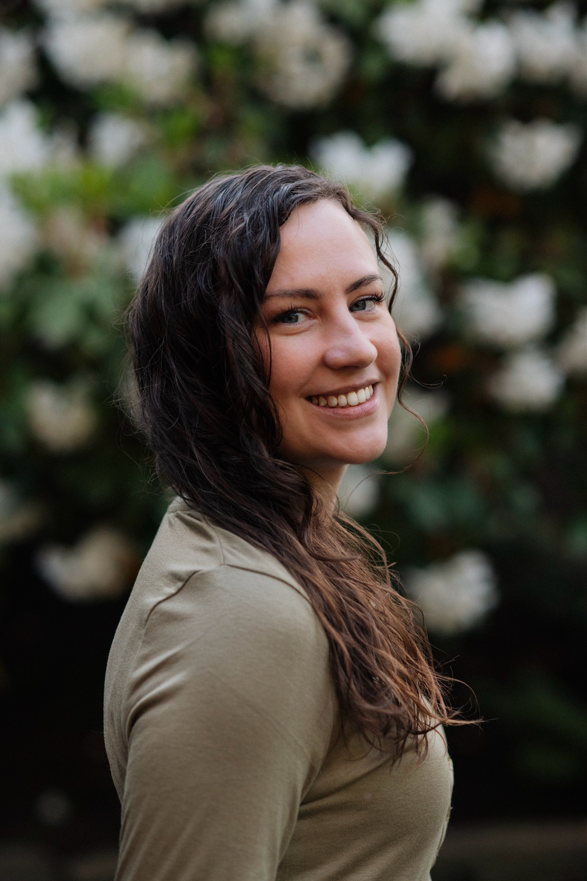 A young woman with long, wavy brown hair smiling outdoors with green foliage and white flowers in the background.