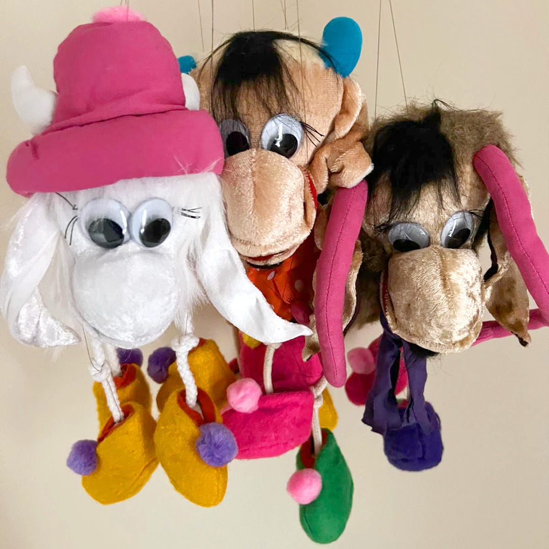 Four plush toys resembling characters with large googly eyes, hanging from a colorful fabric structure against a plain wall background.