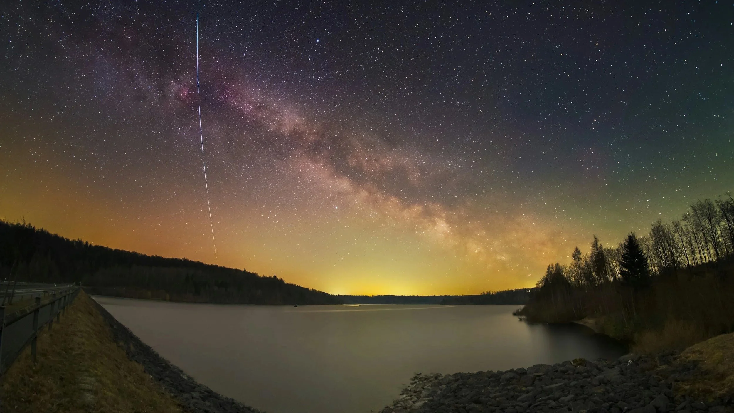 Night sky filled with stars and the Milky Way galaxy over a calm lake, with a treeline on the right and a dam or embankment on the left with a metal railing.